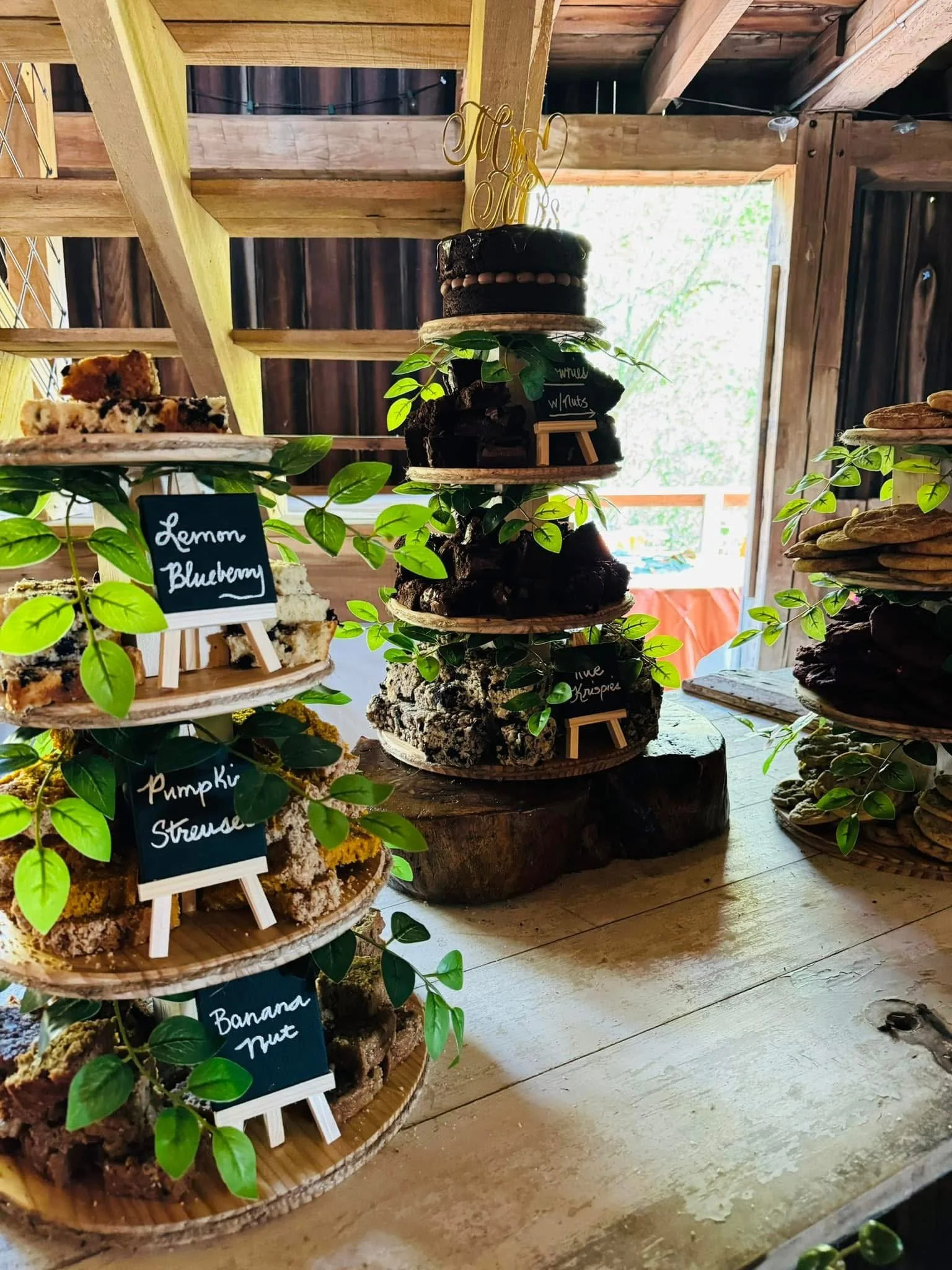 Dessert display with layered cakes on wooden tiered stands and small chalkboard signs labeling flavors like lemon blueberry, pumpkin, banana nut, and apple crumbles inside a rustic wooden structure.