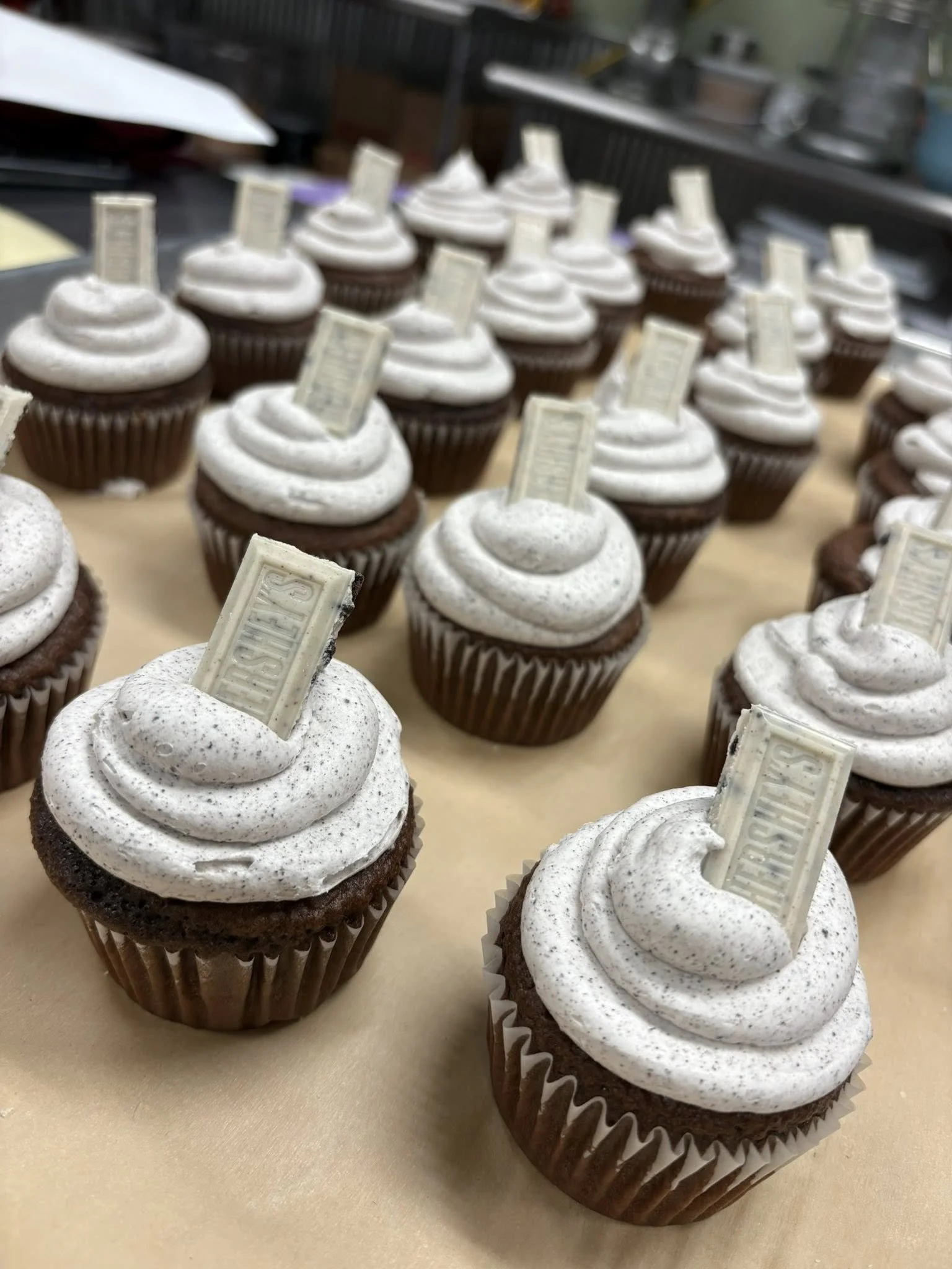 A group of chocolate cupcakes with white frosting topped with a white chocolate tag, arranged on a beige surface in a bakery or kitchen setting.