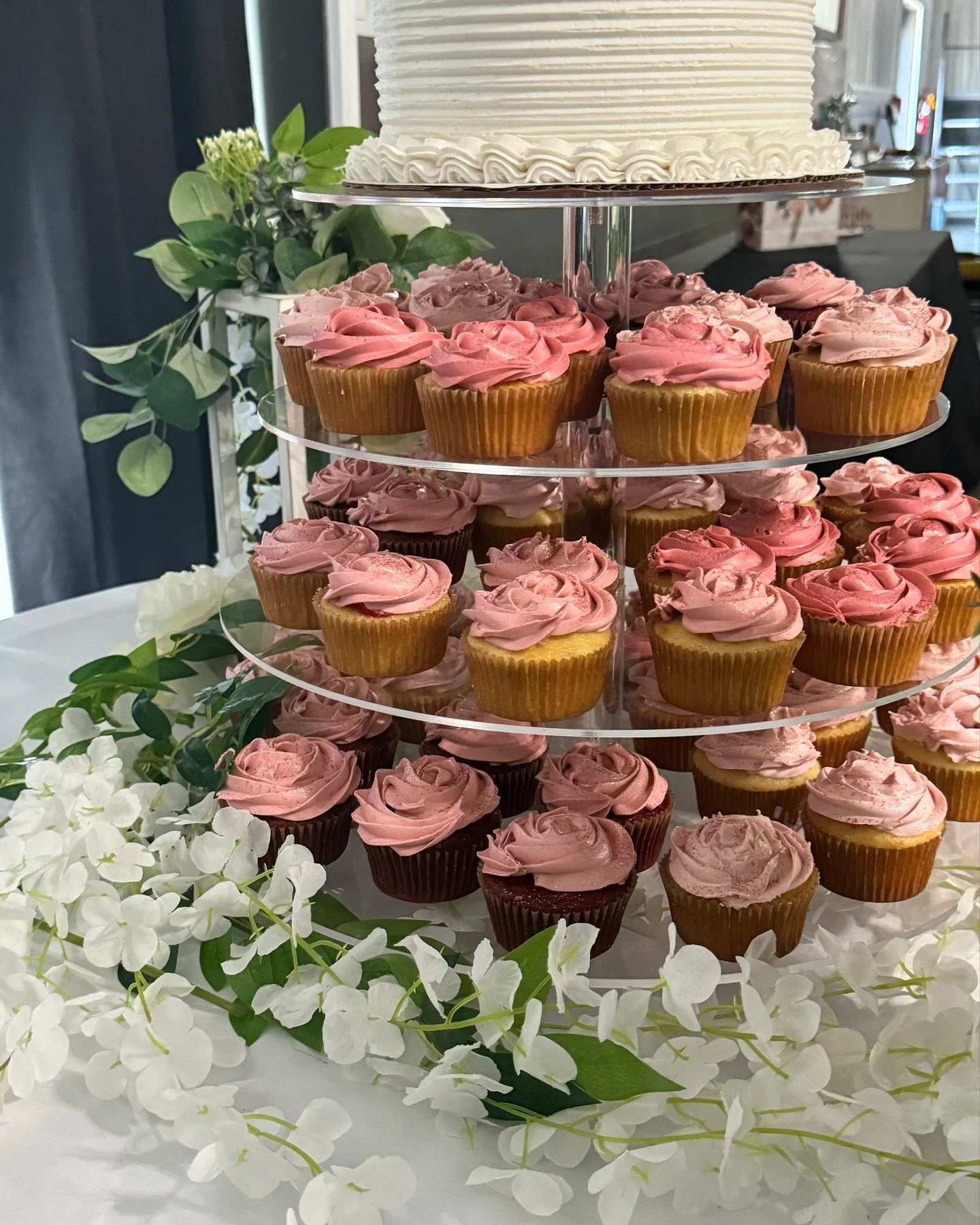 Tiered tray of cupcakes with pink and brown frosting, decorated with white and green floral accents.