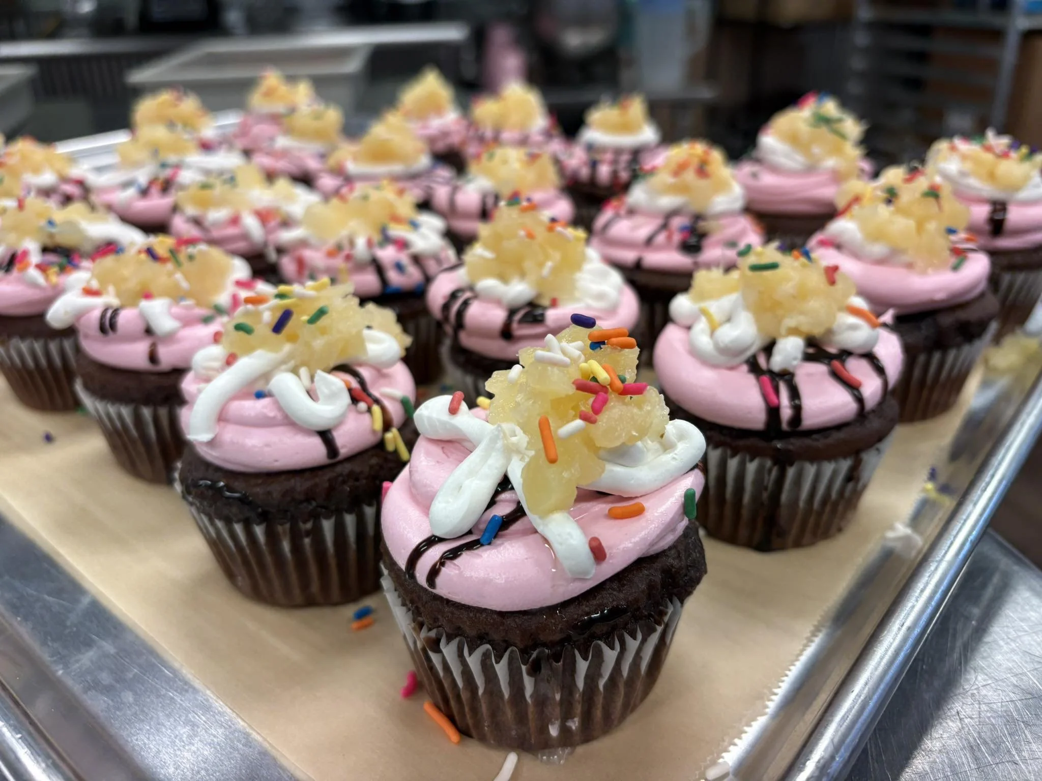A tray of pink frosted cupcakes decorated with colorful sprinkles, white and dark chocolate drizzles, and a pineapple chunk on top.
