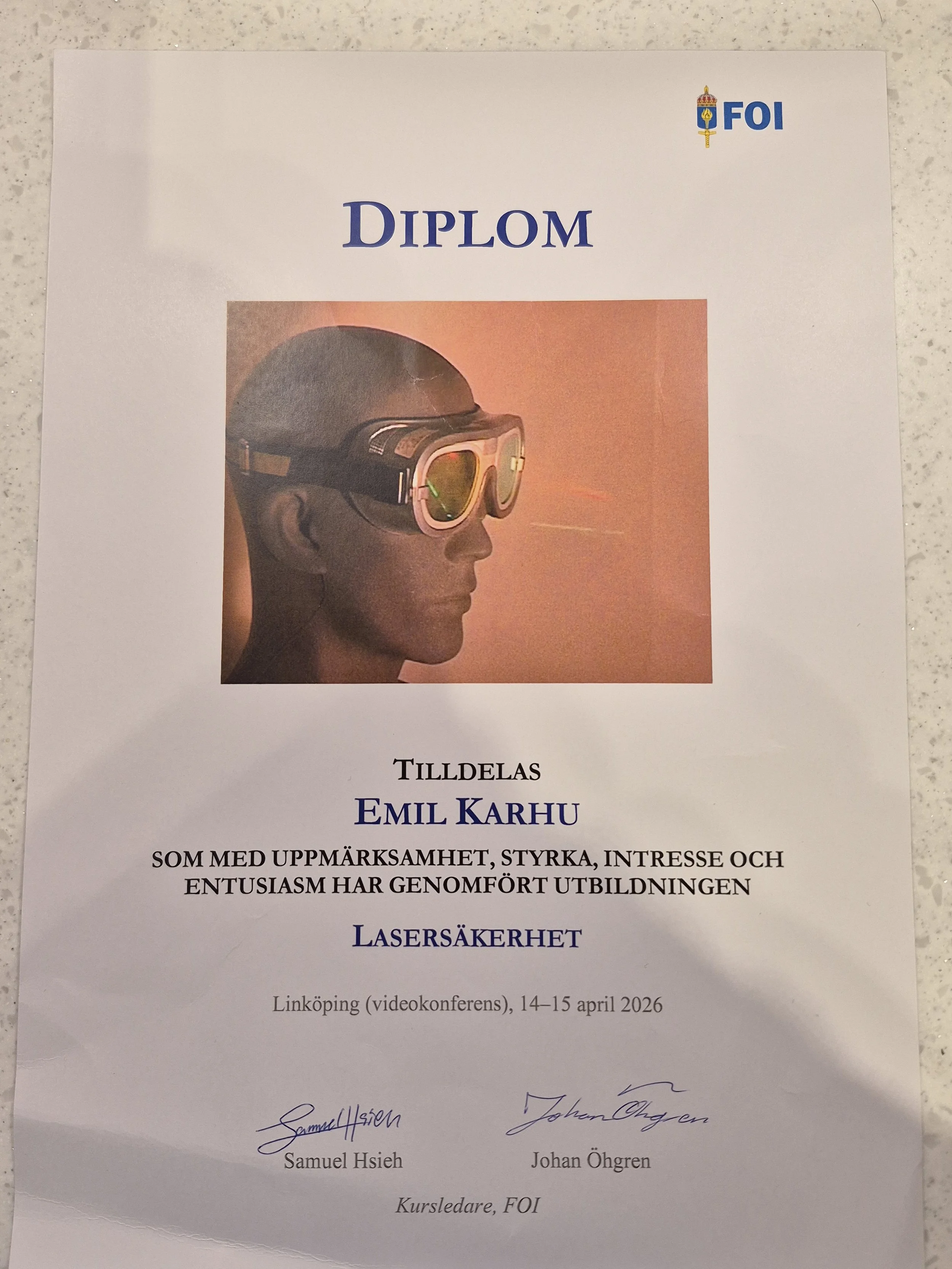 Ett diplom tilldelat Emil Karhu från FOI, med bild av en person med glasögon och skyddsutrustning. Texten nämner lasersäkerhet och videokonferens den 14-15 april 2026 i Linköping.
