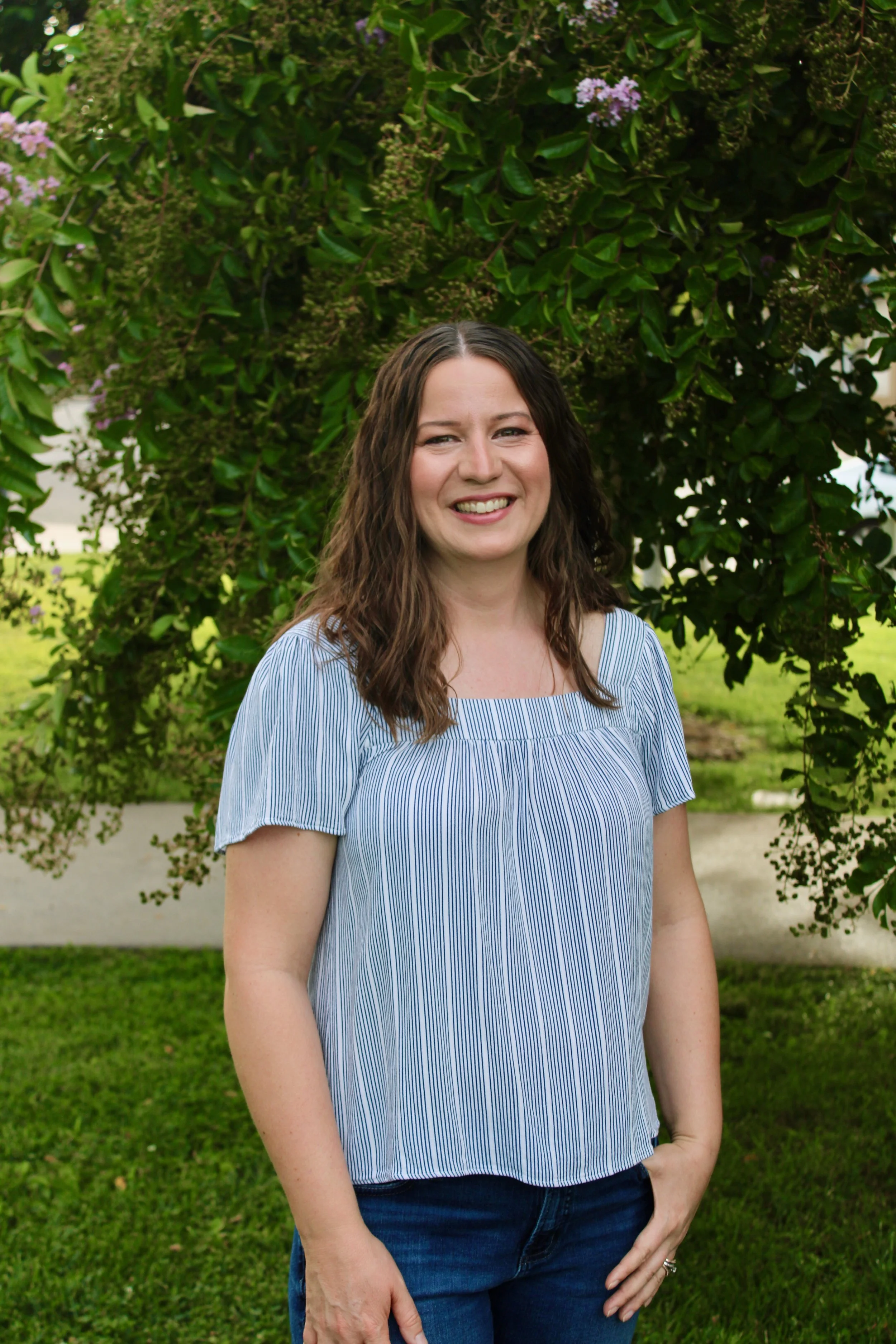 A woman with brown hair smiling, standing outdoors in front of a large green shrub with purple flowers, wearing a blue and white striped top and blue jeans.