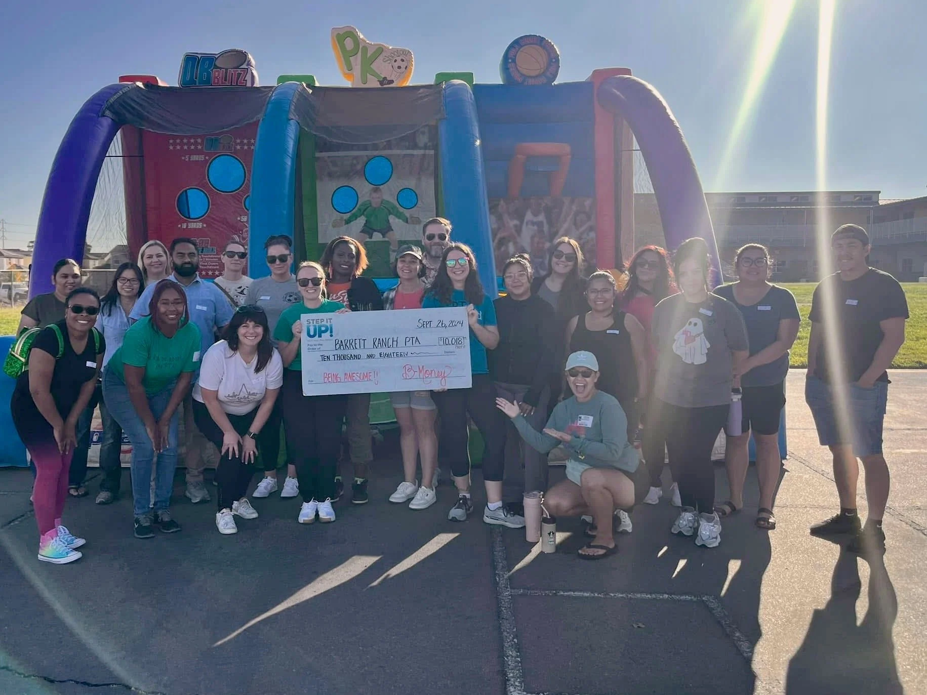 A group of diverse adults posing outdoors with a large check for $10,018 made out to Barret Ranch PTA, surrounded by inflatable structures and banners, under a bright blue sky.