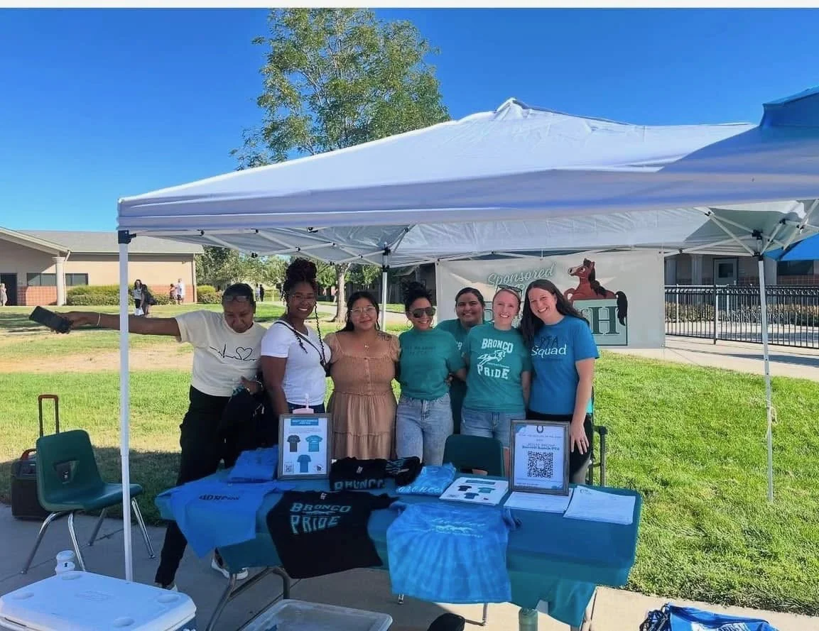 Group of six young women standing under a white tent at an outdoor event, smiling and posing for the photo. The table in front of them displays various T-shirts with slogans like "Bronco Pride." The background shows a grassy area, trees, and a building.