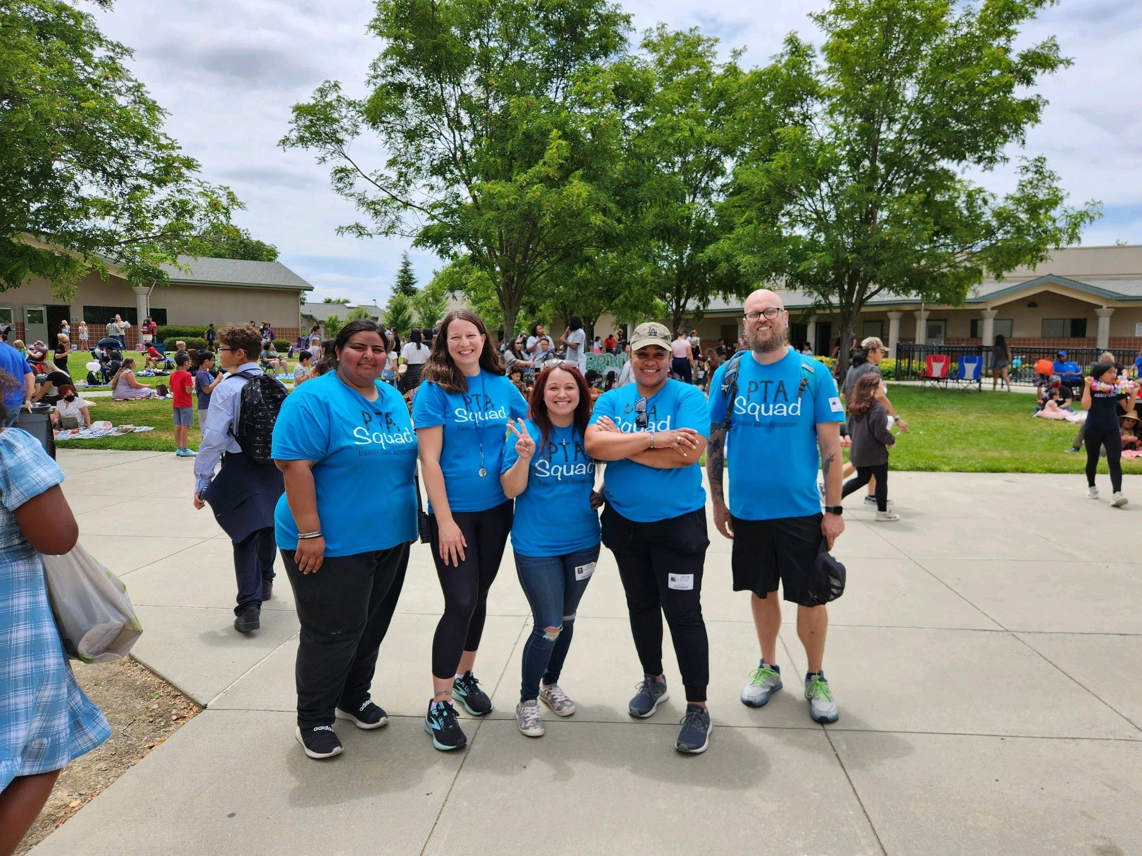 Group of five diverse people smiling and posing together outdoors during an event, with many other people in the background, a building, trees, and a cloudy sky.
