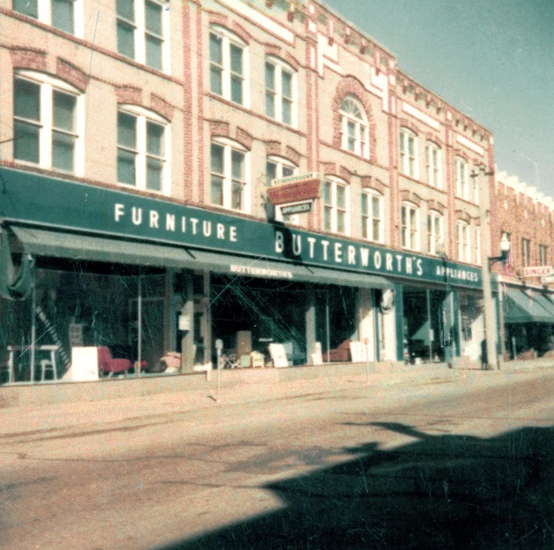 A faded image of the facade of Butterworth's Furniture Store, with broad glass display windows lining a sidewalk  – genealogy research. © Butterworth Research