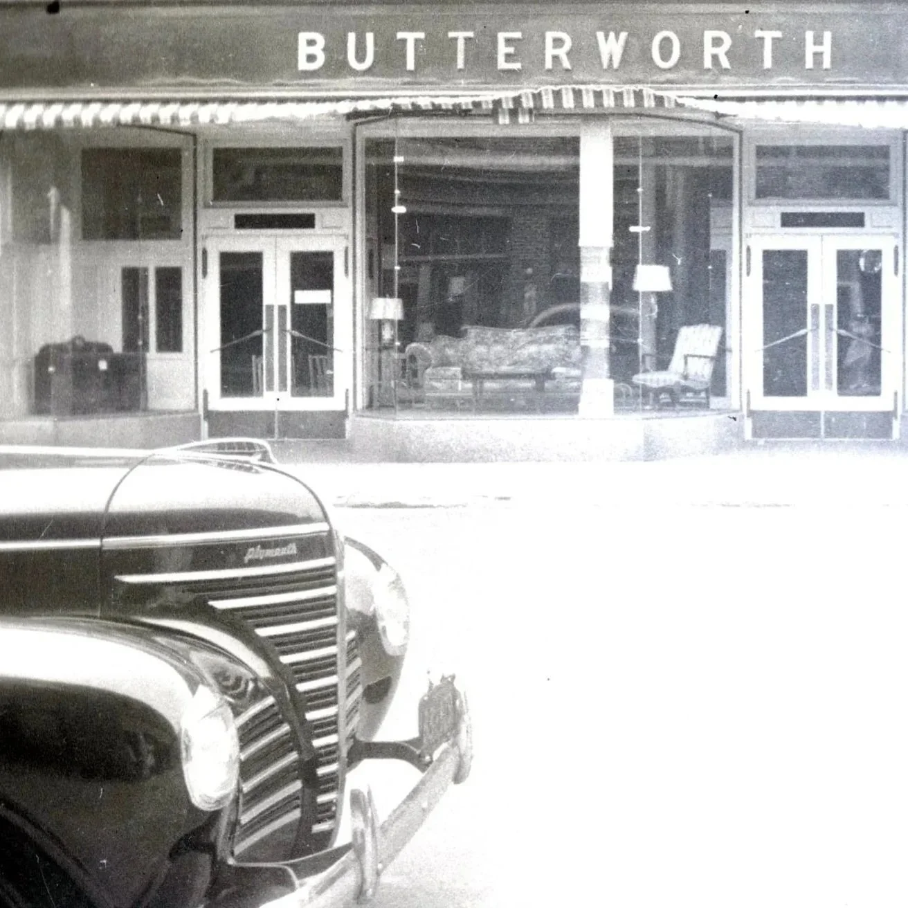 A black and white image of the facade of Butterworth Furniture Store with an antique Plymouth car entering the frame from the left  – genealogy research. © Butterworth Research