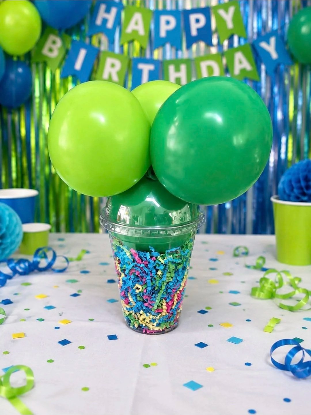 Green and blue balloons arranged in a cup filled with colorful shredded paper, with a birthday party decoration in the background and scattered confetti on the table.