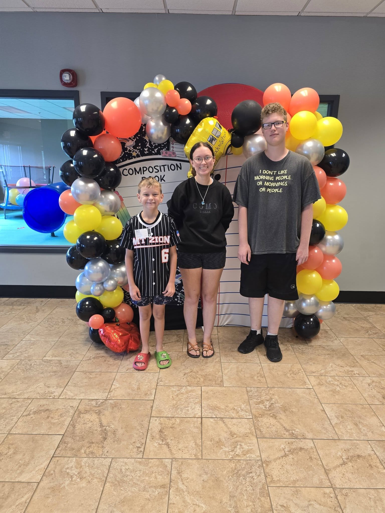 Three people posing in front of a colorful balloon arch at a school event, with a sign that reads 'Composition Book' in the background. The group includes a young boy wearing a sports jersey and slippers, a woman in casual clothing, and a tall young 