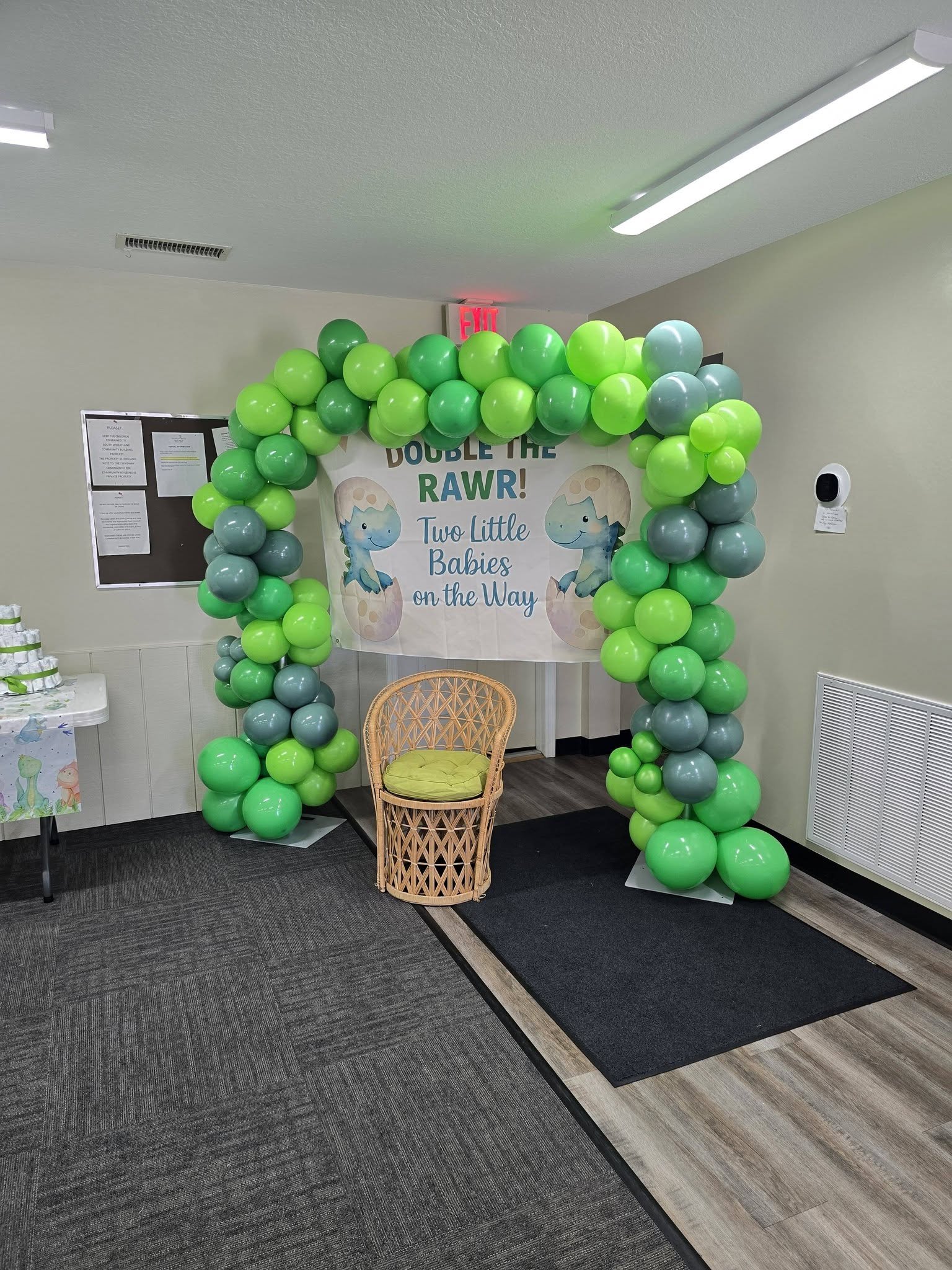 Decorative balloon arch with green and gray balloons at a baby shower, with a sign that reads 'Double the RAWR! Two Little Babies on the Way' featuring cartoon dinosaur illustrations.