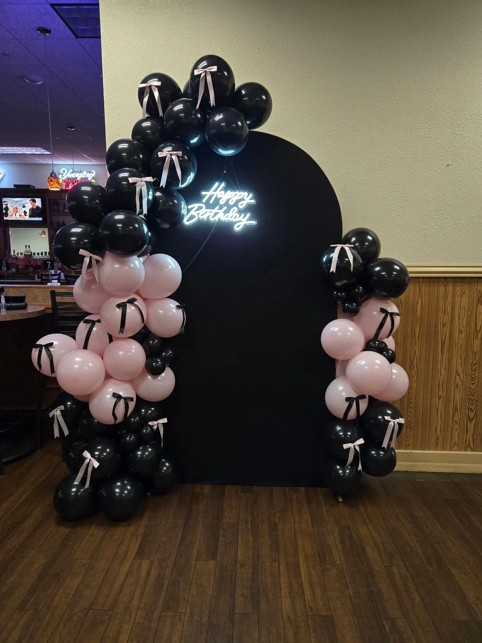 Black and pink balloons arranged in an arch with a black backdrop that has a neon sign reading 'Happy Birthday' in white letters, decorated with pink ribbons tying some of the balloons.