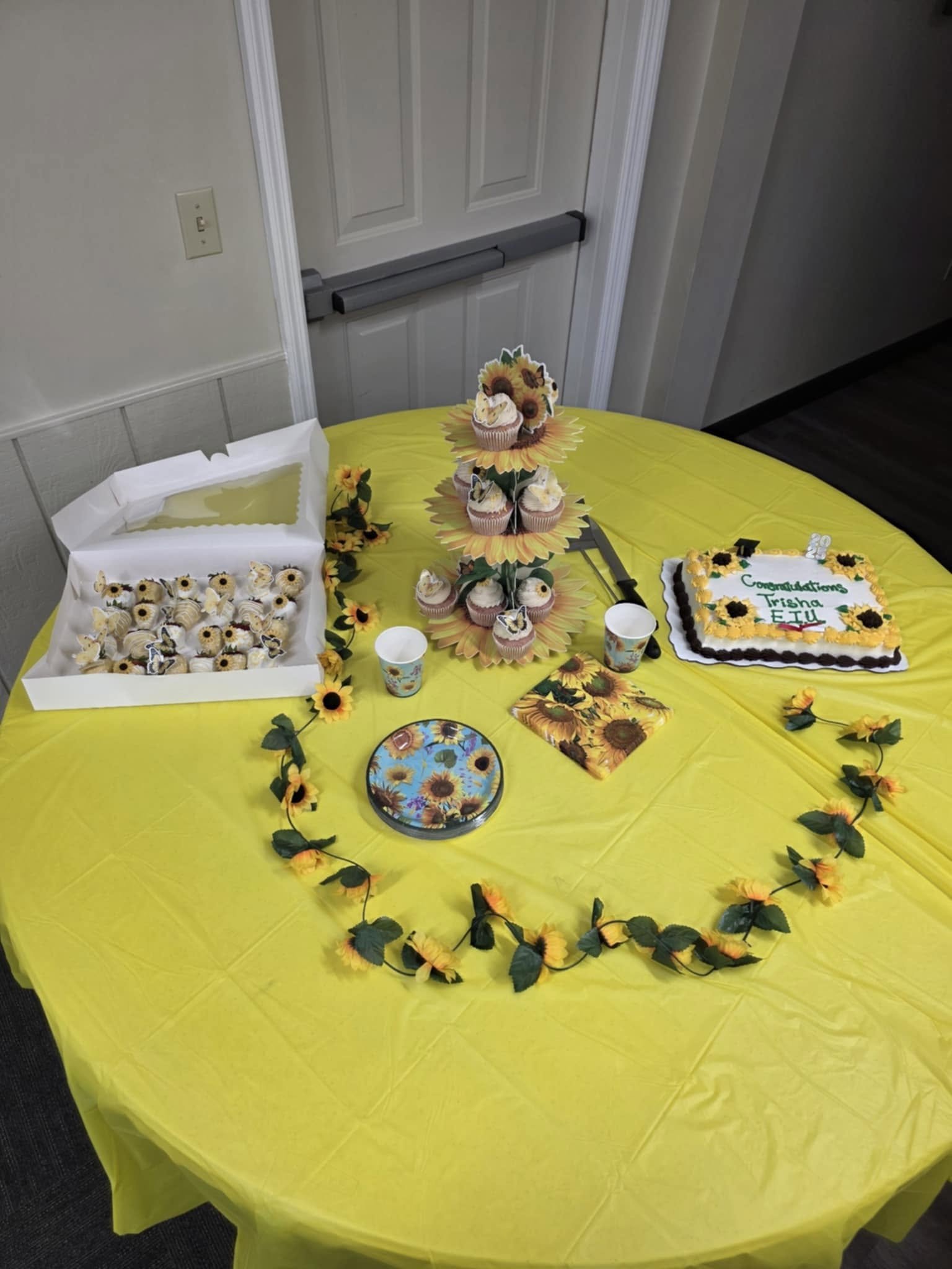 A decorated table with yellow sunflower-themed party supplies, cupcakes with sunflower toppers, sunflower garland, sunflower-themed napkins and plates, a congratulatory cake with sunflower decoration, and sunflower-themed cups, set against a plain wa