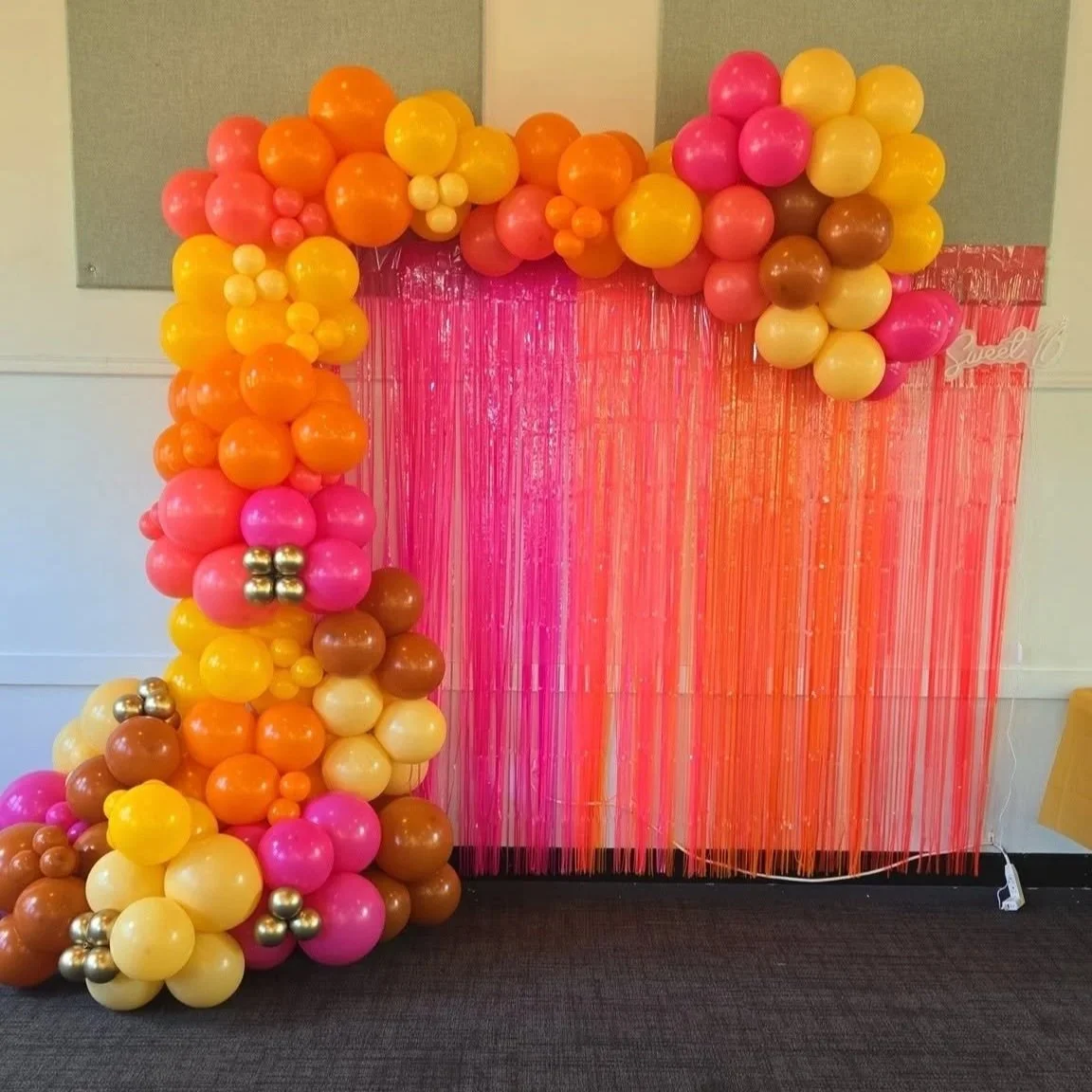 Decorative balloon arch with pink, yellow, orange, brown, and metallic gold balloons next to a pink string curtain backdrop for a celebration.