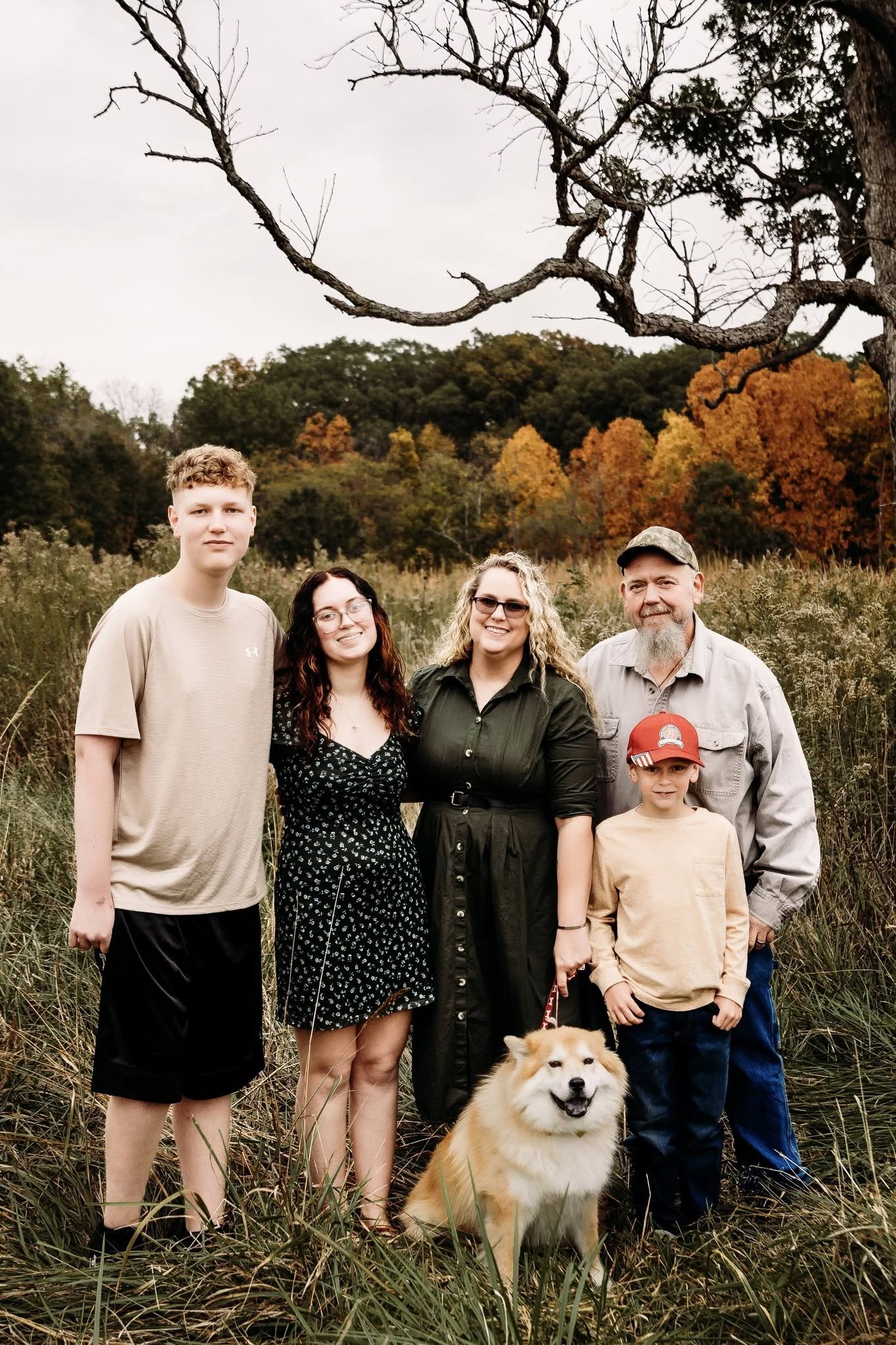 A family of seven people and a dog standing outdoors in a grassy field with trees in the background. The scene appears to be in autumn, with some trees showing orange and yellow leaves and others being leafless, on a cloudy day.