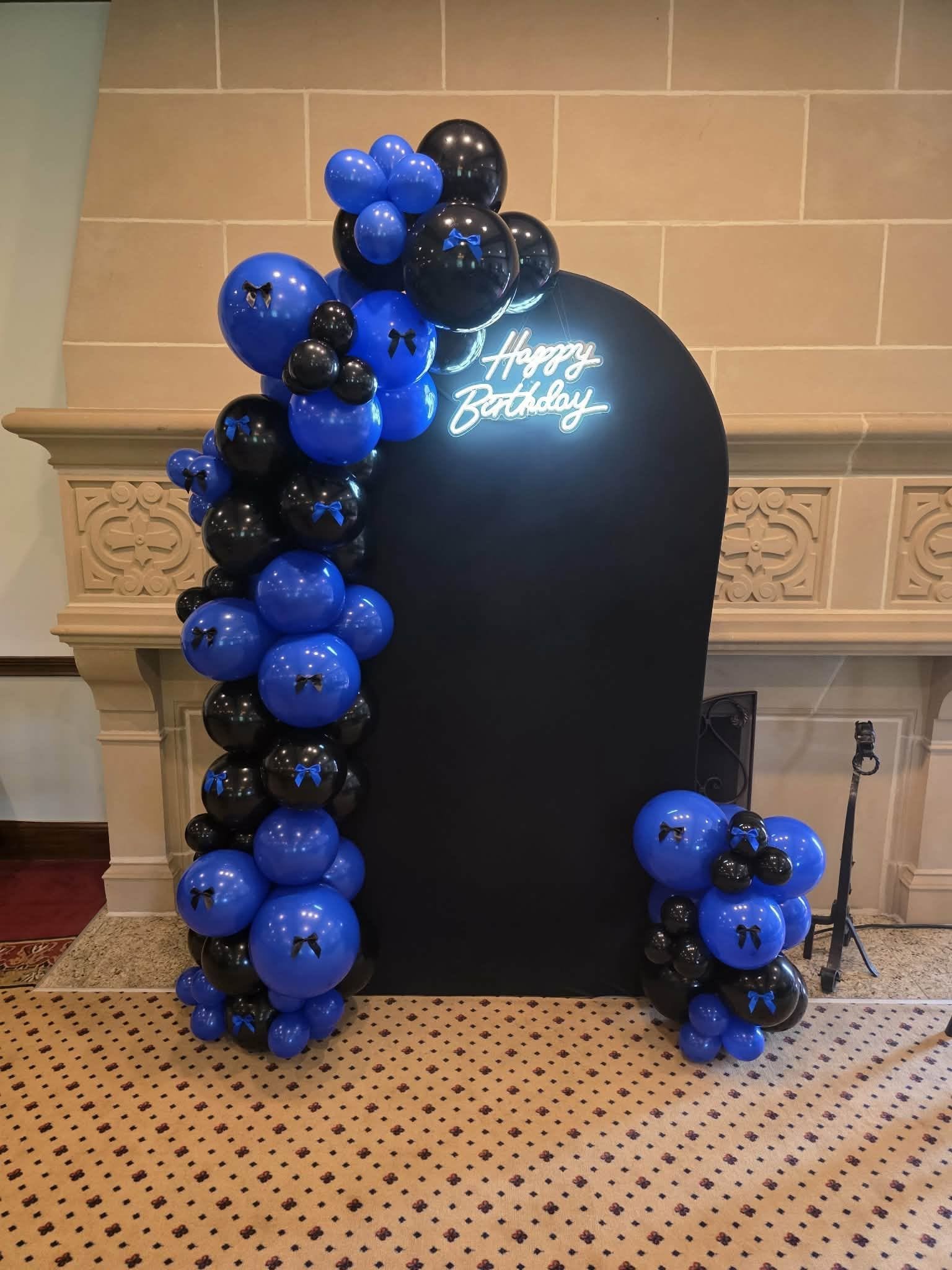 A balloon arch with blue and black balloons arranged in columns around a black backdrop with a white neon sign that says 'Happy Birthday' inside a room with a beige and brown patterned carpet and a fireplace with decorative woodwork.