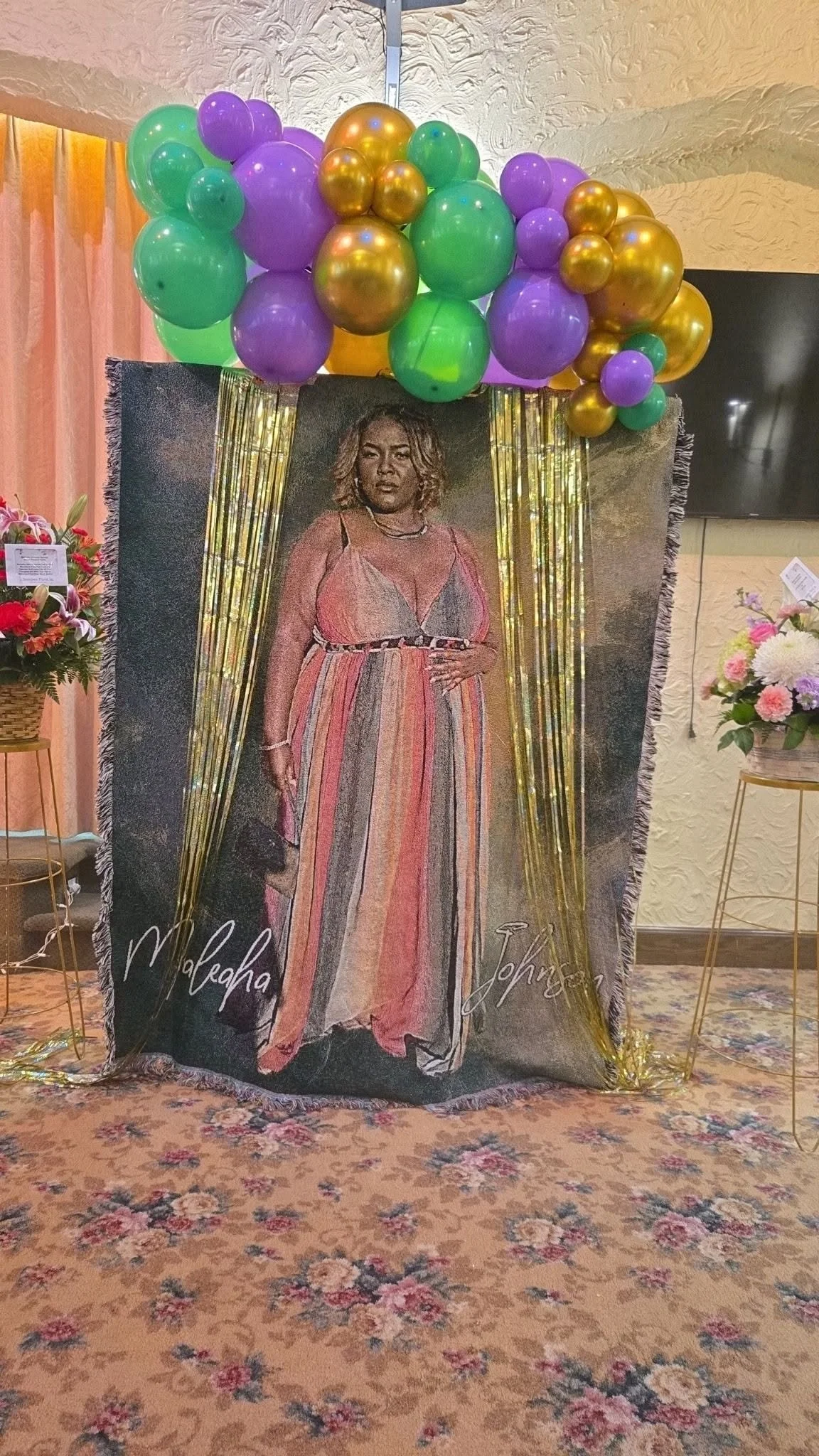 A decorative display featuring a large portrait of a woman with shoulder-length hair, wearing a pink and multicolored dress. The portrait is embellished with black and gold accents and is decorated with gold and purple balloons arranged above it. The