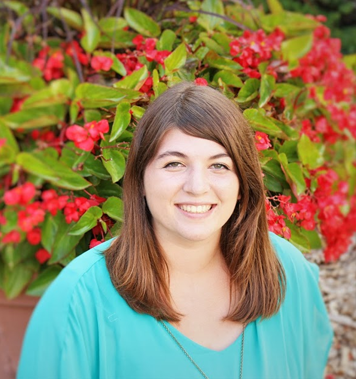 Headshot of author Allison Mulder. She is a young woman with hazel eyes and chestnut brown hair that falls just past her shoulders. She is wearing a bright cyan blouse and is sitting in front of a large bush with red flowers.