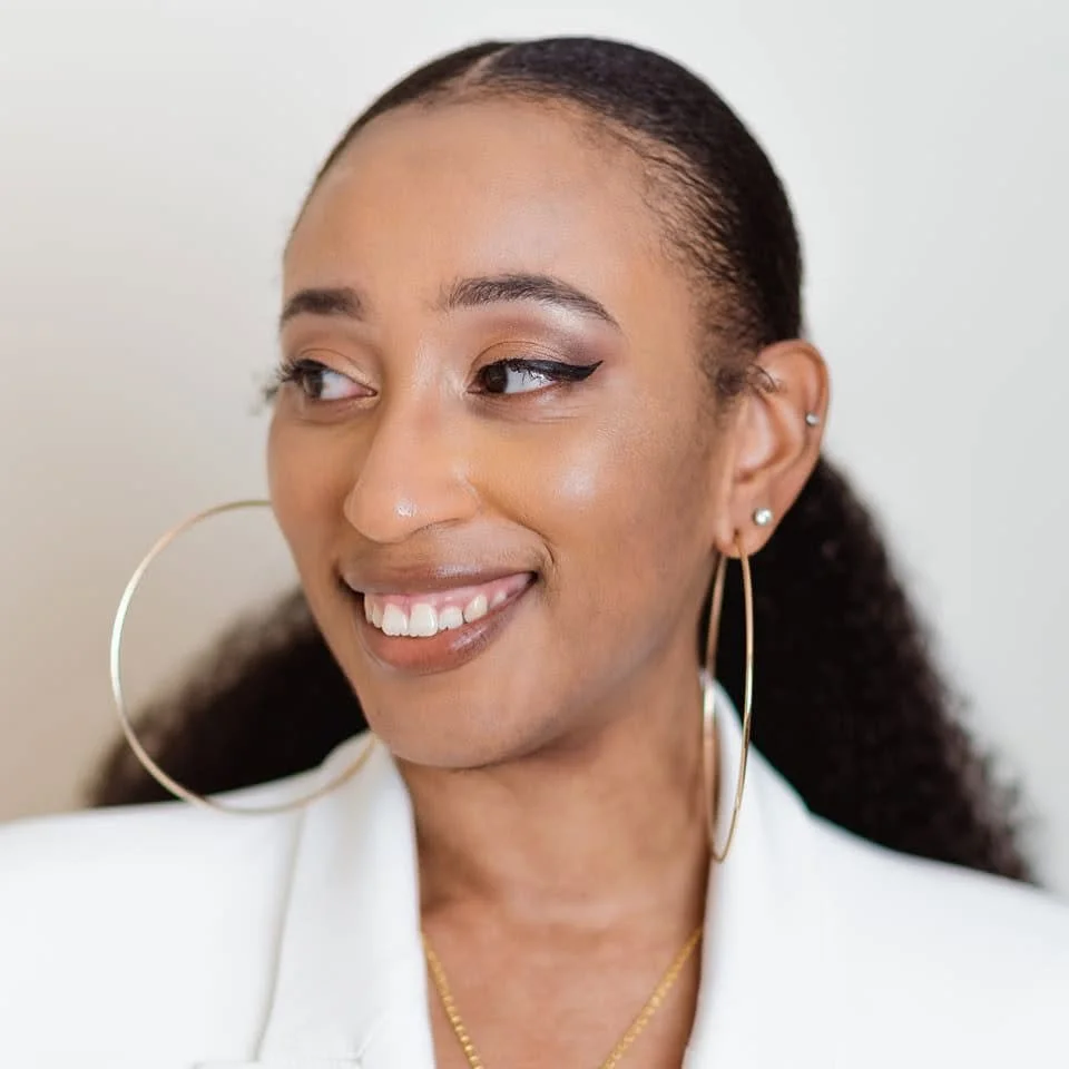 Zanaiya Leon, CEO & Found of RICHVLLE. Description: A Black woman smiling wearing a white blazer, large hoop earrings and gold necklace, wearing makeup, and looking to her left.