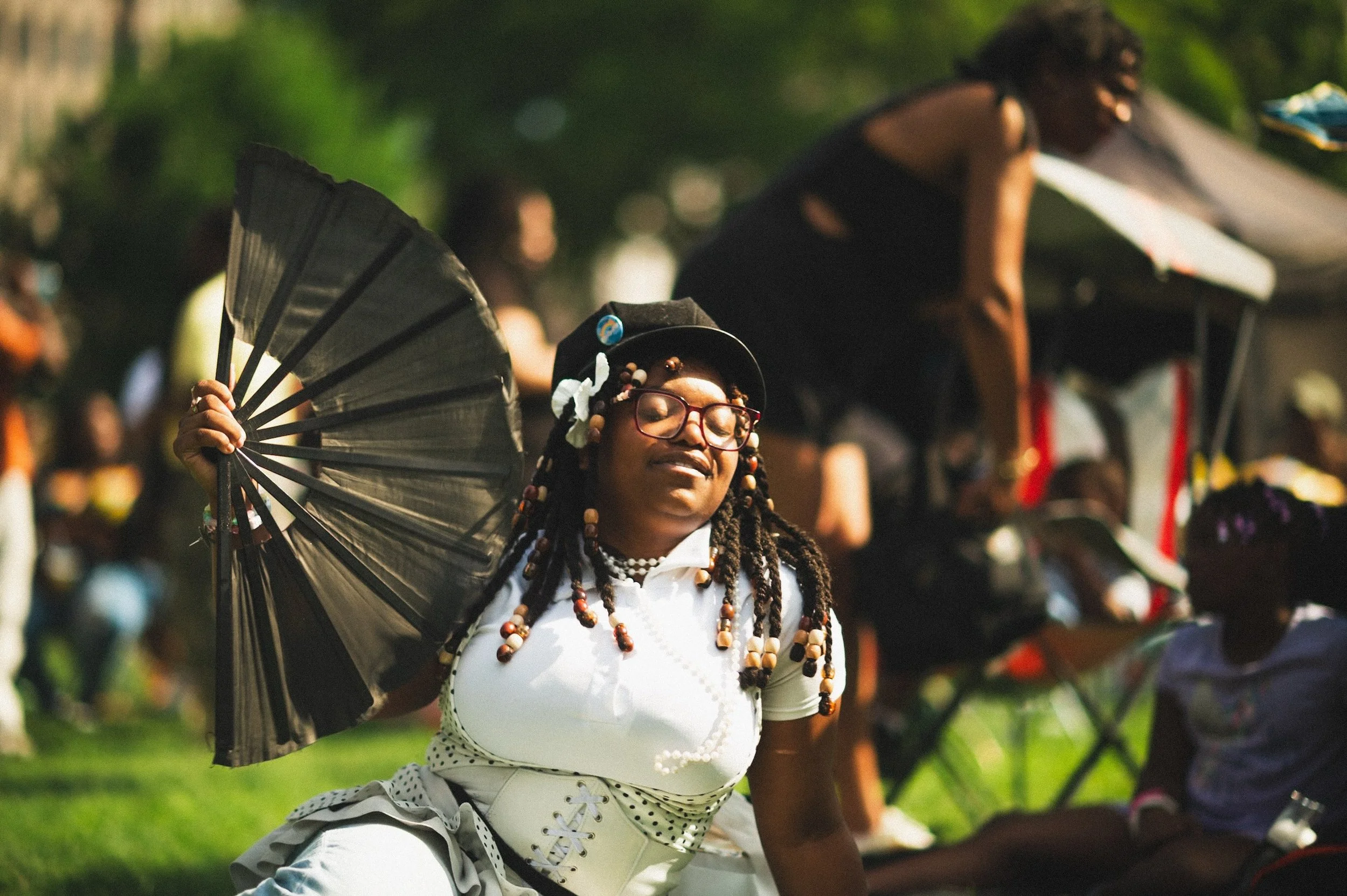 A woman with glasses and braided hair adorned with beads, dressed in a white outfit with polka dots, holding a black fan with a smile and eyes closed during the New Haven, CT 2026 Black Wall Street celebration with blurred people in the background.