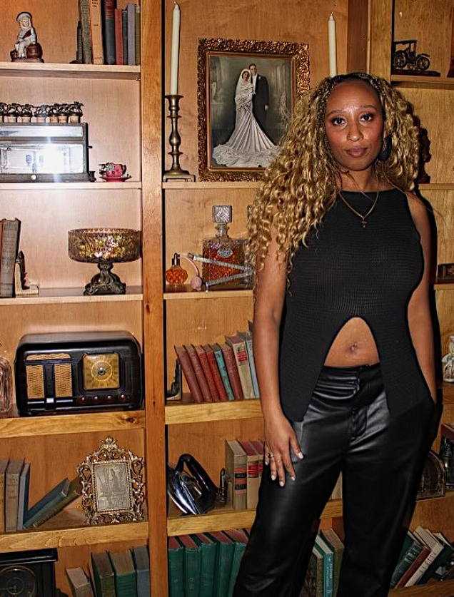 Zanaiya Leon - Woman with curly blonde hair wearing a black sleeveless top and black leather pants standing in front of wooden bookshelves filled with books and decorative items. There is a framed wedding photo and various ornaments on the shelves.