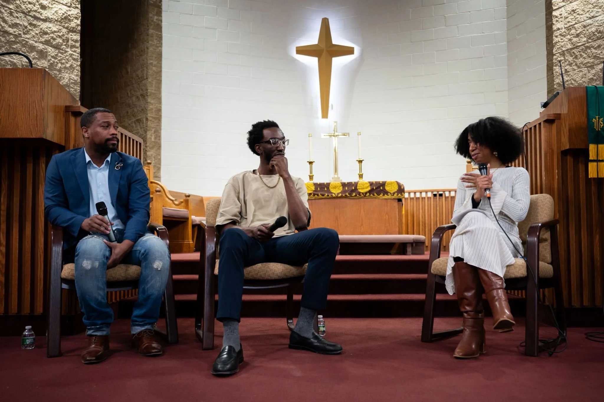Three people sitting on chairs in a church setting, with a woman on the right holding a microphone and speaking. A man on the left holds a microphone, and another man in the middle has his hand on his chin. The background features a white wall with a gold cross and altar behind them, and two wooden stands on either side.