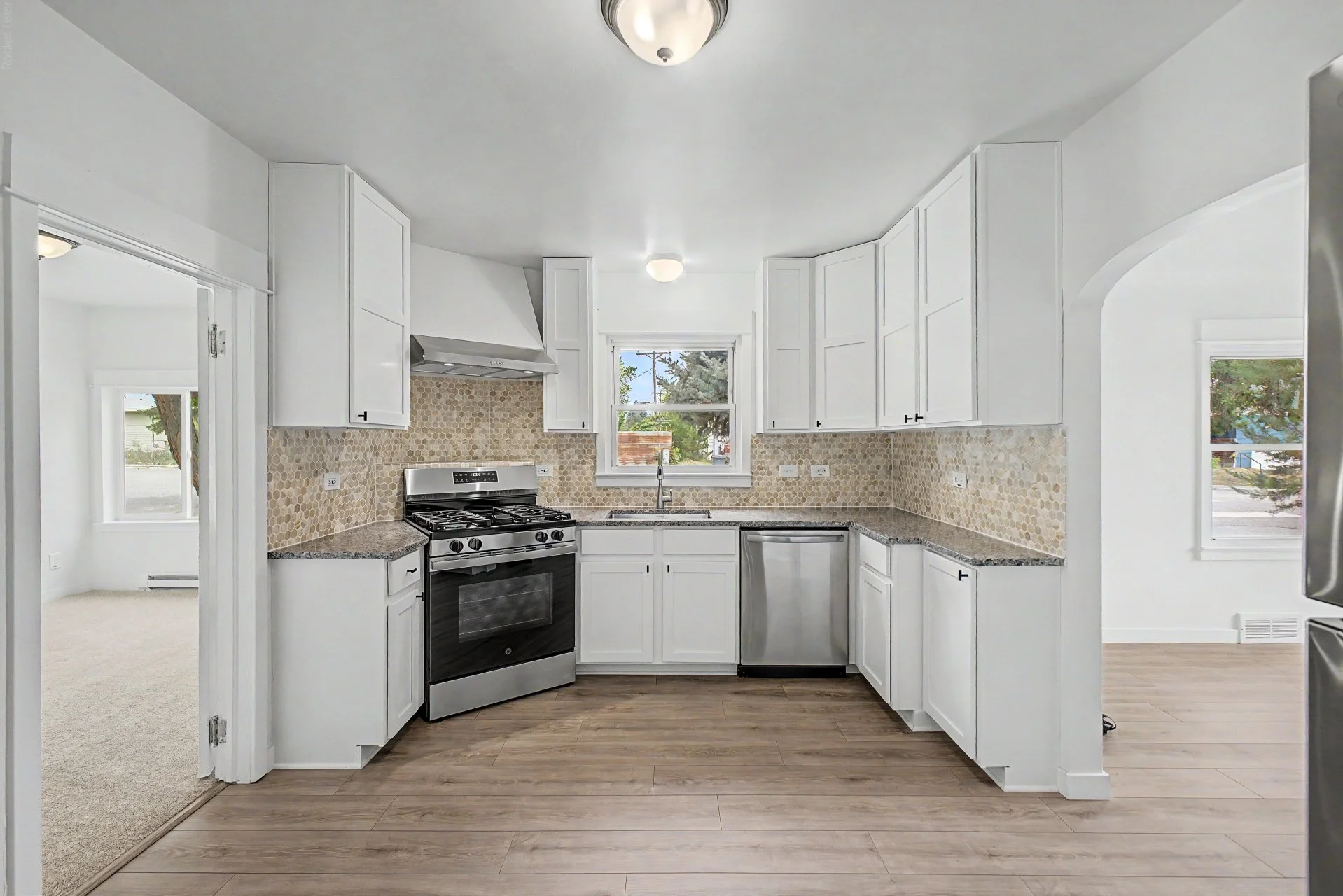 Brand new white kitchen with granite countertops, beige hexagonal tile backsplash, stainless steel stove, dishwasher, and oven hood. There are cabinets above and below the countertops, with a window above the sink showing trees outside.