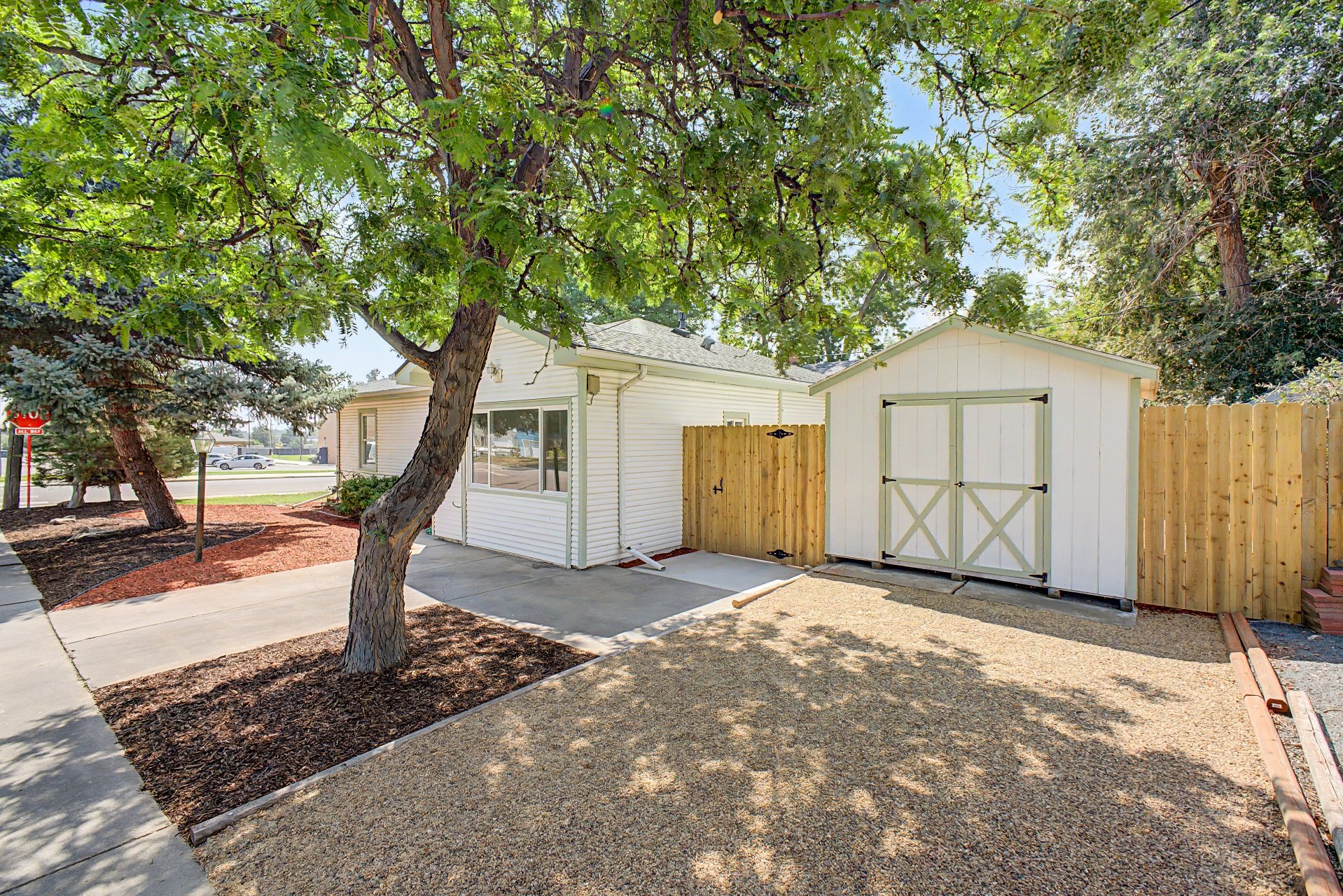 Freshly landscaped front yard of a house with a large tree, white house with sliding window, concrete patio, small white shed with black hinges, wooden privacy fence, and landscaped area with mulch and gravel.