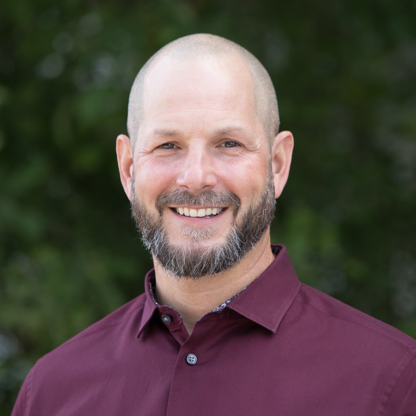 A headshot of the author, smiling with a beard and bald head wearing a maroon collared shirt, standing outdoors with a blurred green background.
