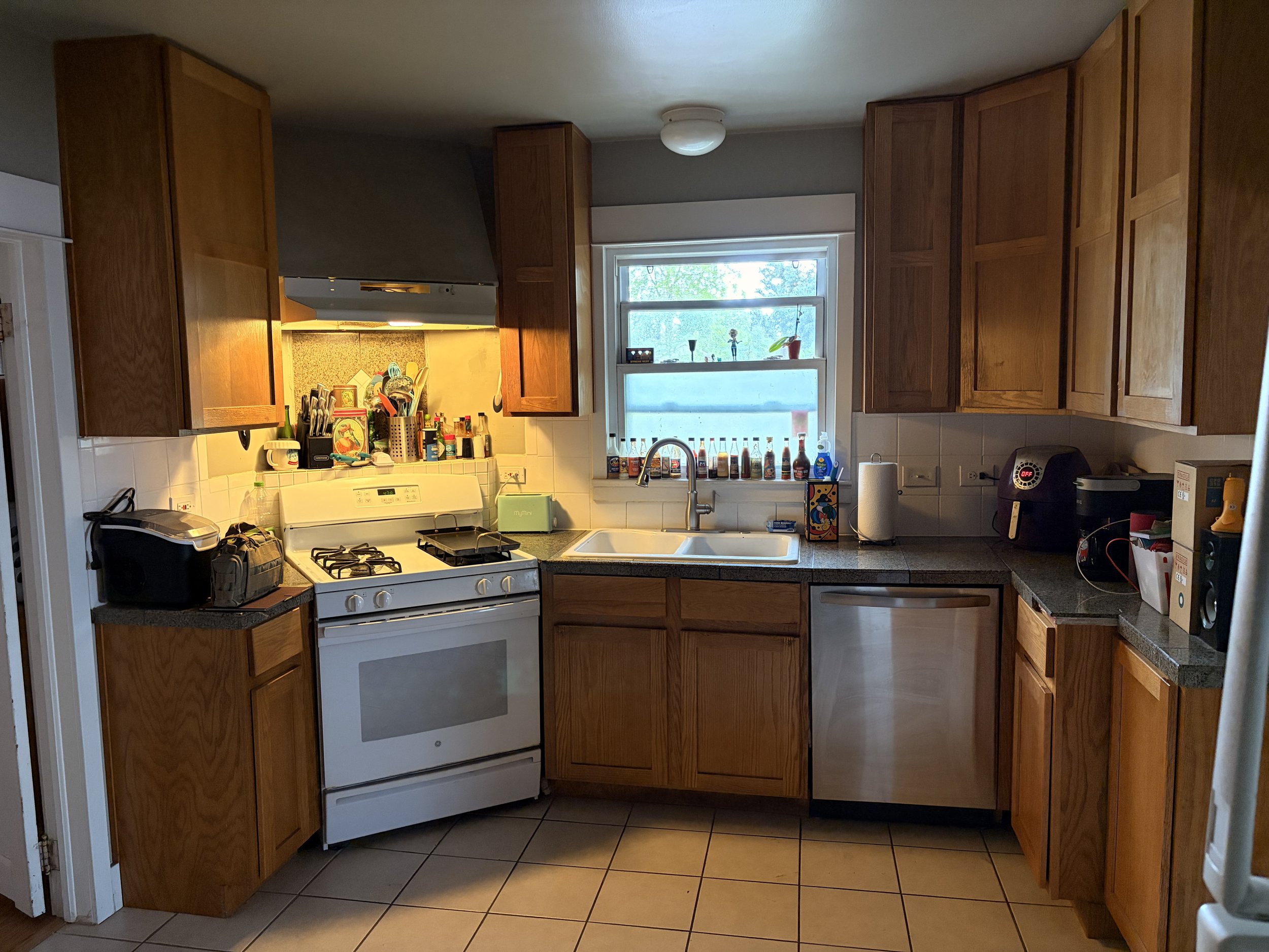 An old dirty kitchen in need of repair with wooden cabinets, a white stove, a window above the sink, and various bottles and kitchen items on the counter and windowsill.