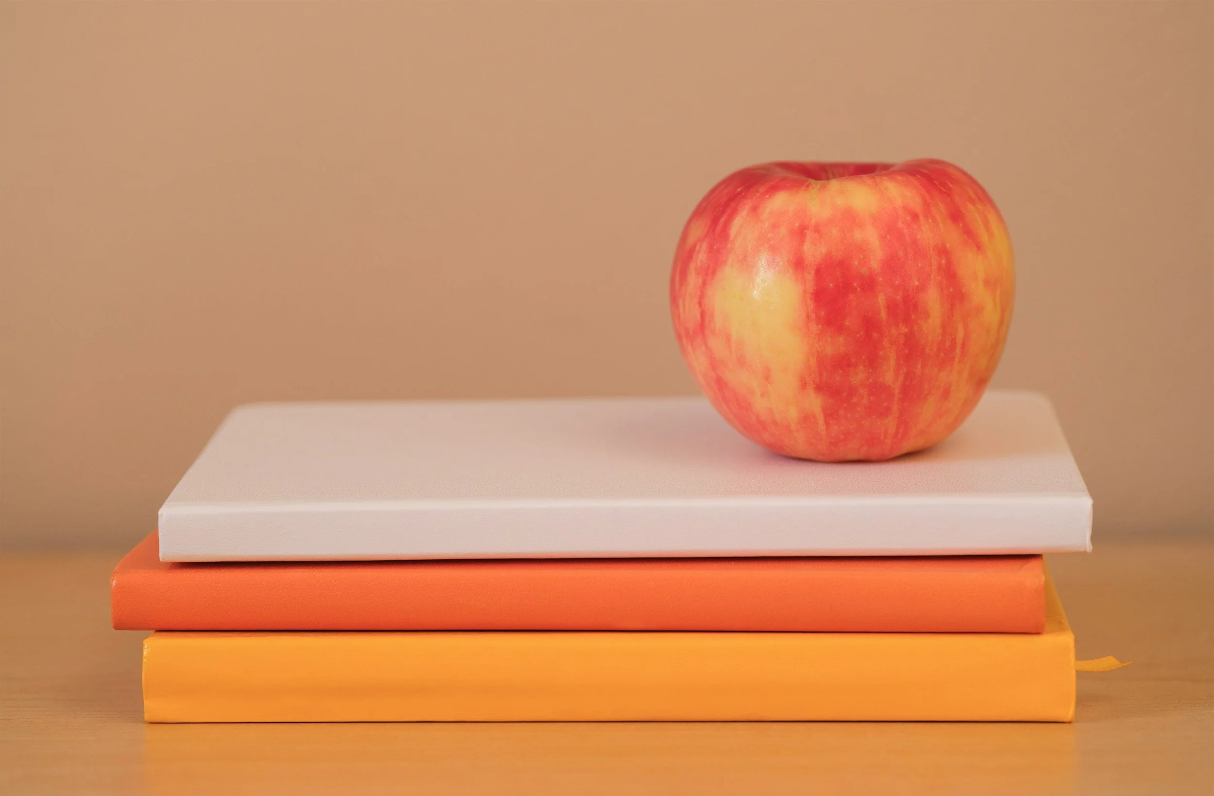 A stack of four colored books with a red and yellow apple on top, against a beige background.