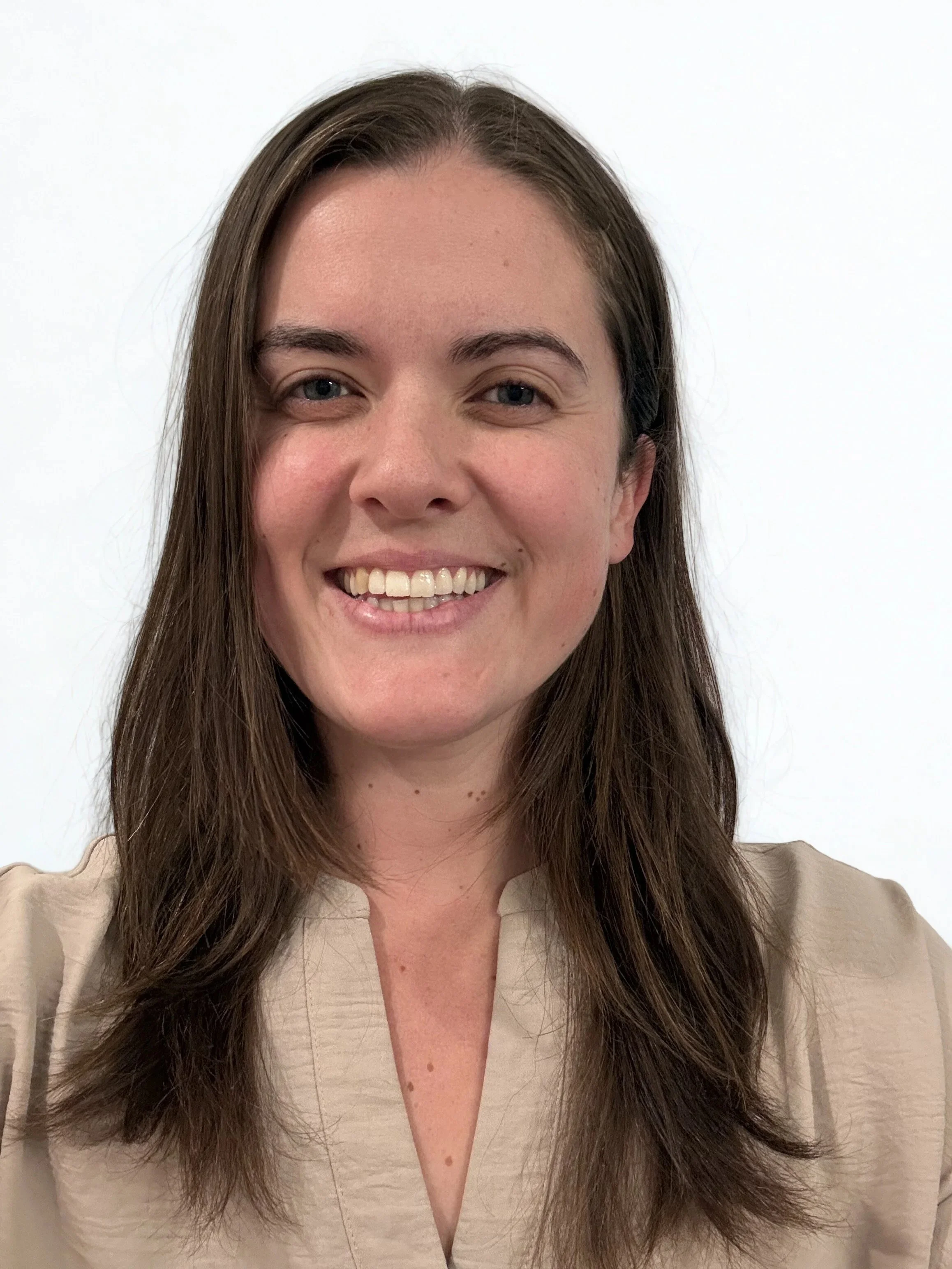 A smiling woman with long brown hair, blue eyes, and light skin, wearing a beige top, standing against a plain white background.