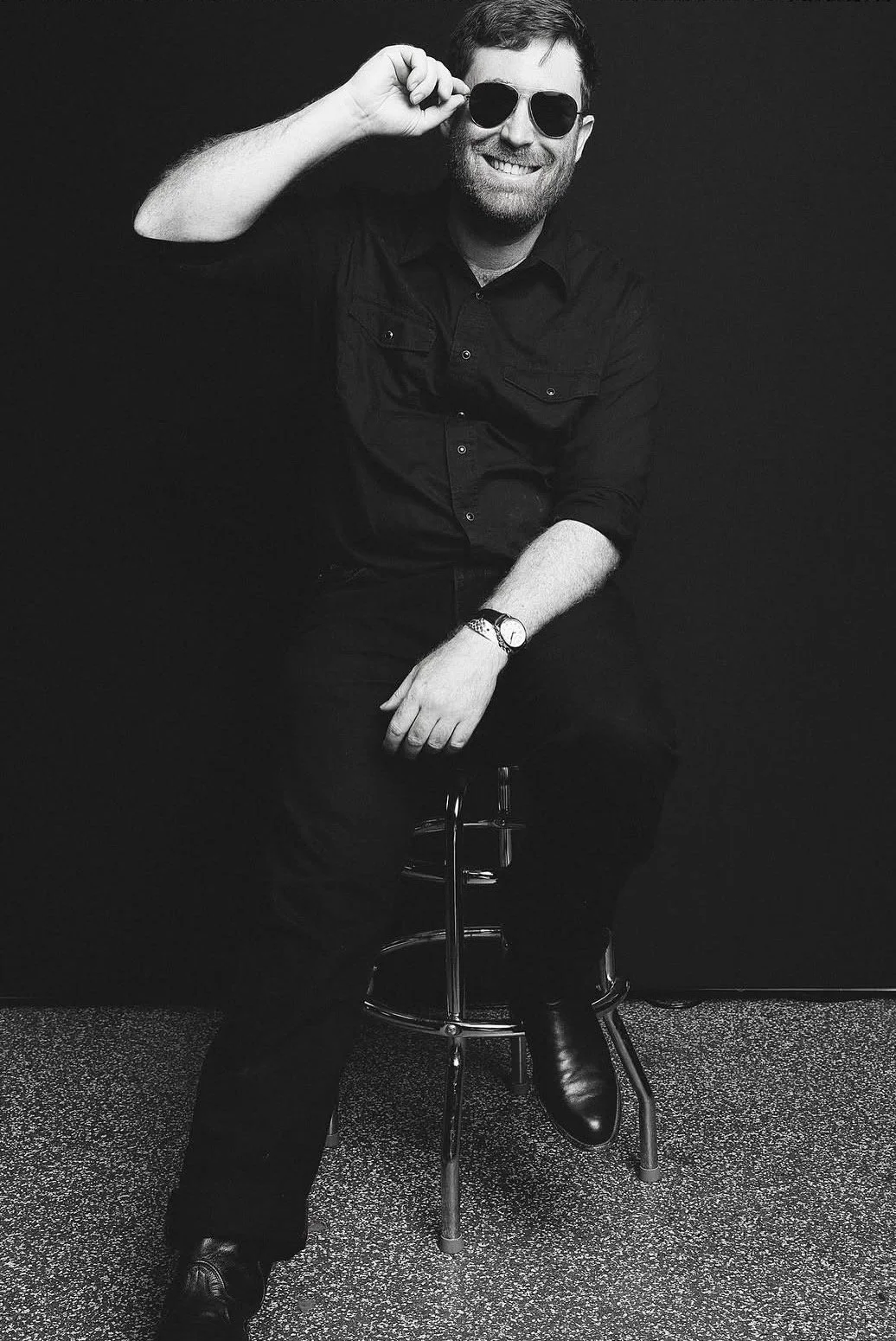 Black and white photo of a smiling man wearing sunglasses, a dark button-up shirt, a wristwatch, and black pants. He is sitting on a stool against a dark background and holding his sunglasses with his right hand.