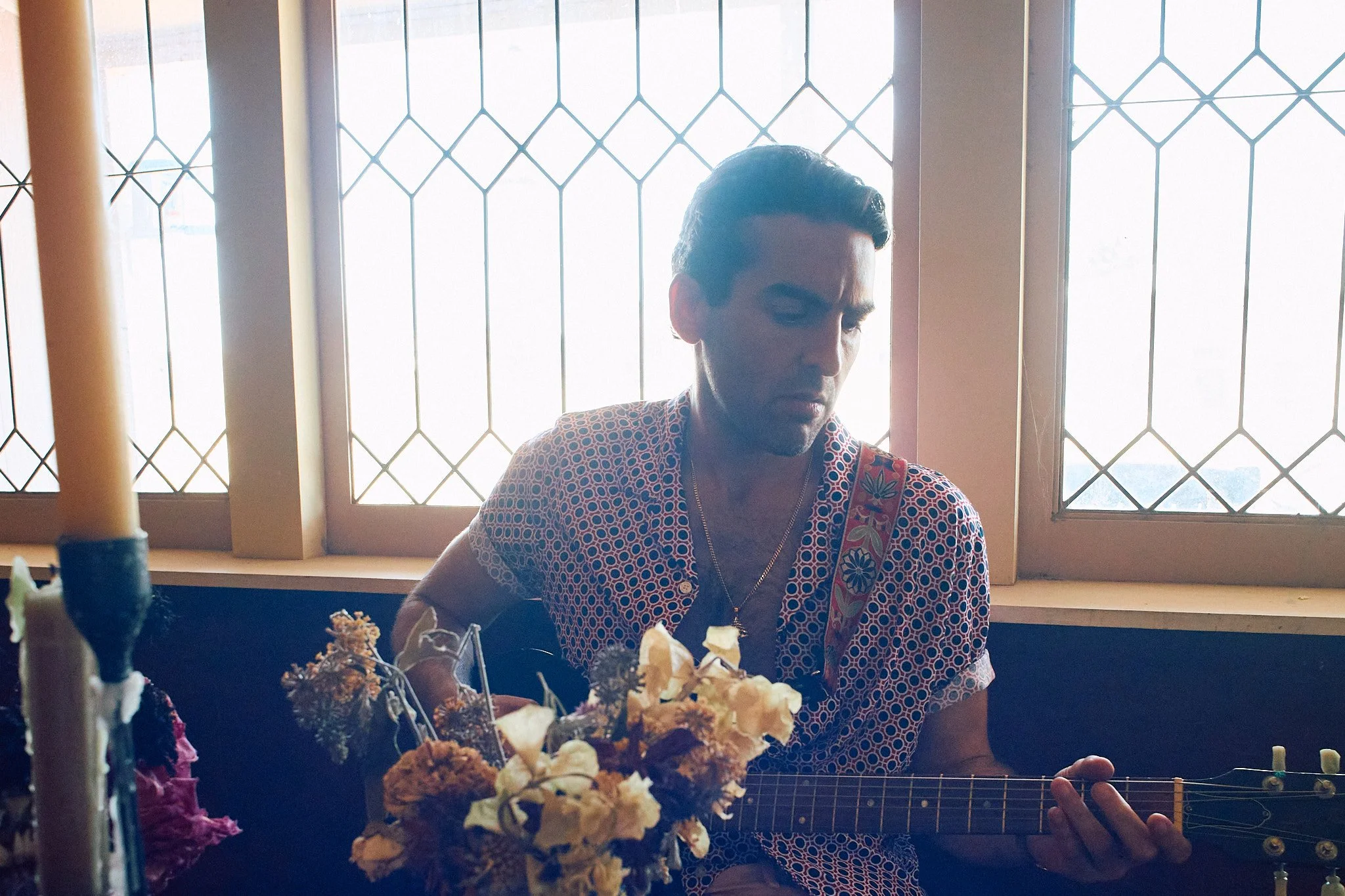 A young man playing an acoustic guitar behind a table with dried flowers, sitting in front of a window with decorative metal bars, during daytime.