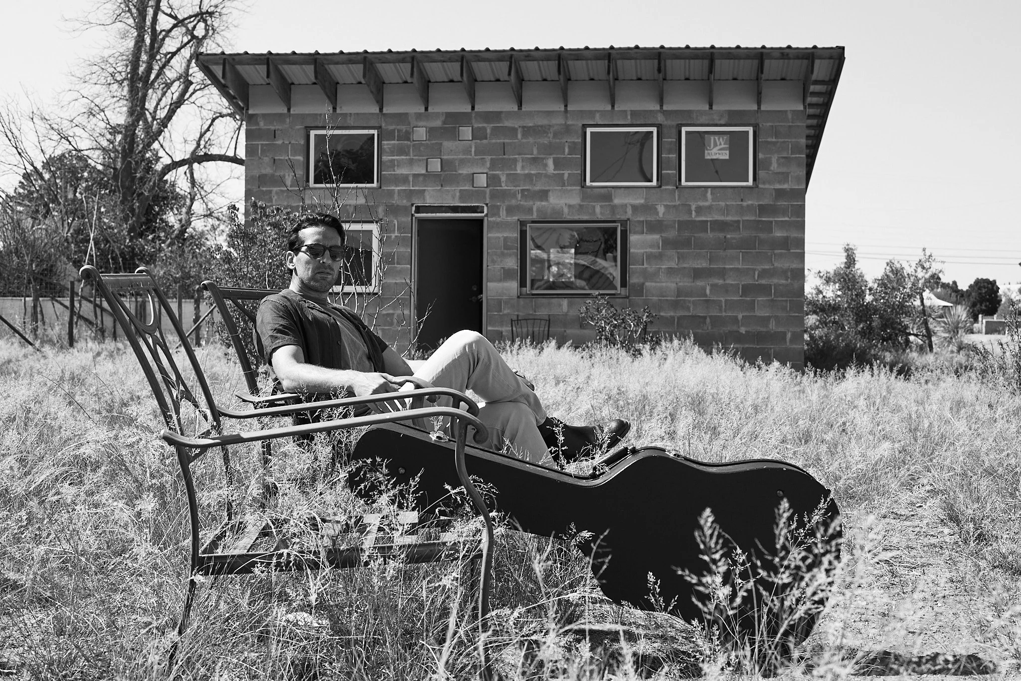 Black and white photo of a man sitting in a wheelchair outdoors in front of a house with brick walls and multiple windows, surrounded by tall grass and trees.
