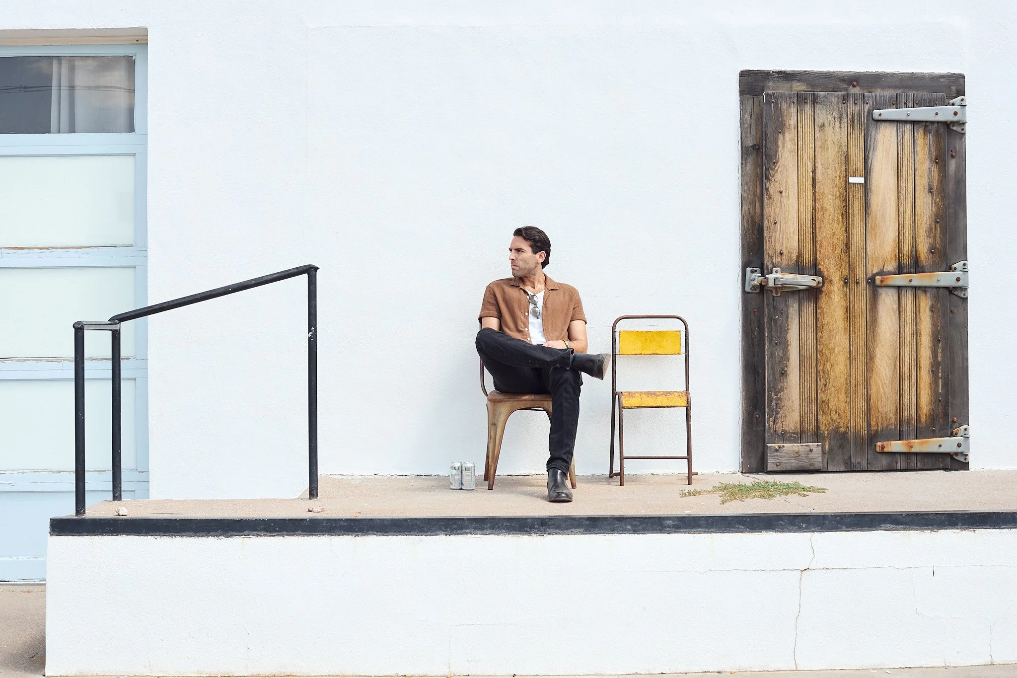 A man sits on a brown chair outside a white building with a large wooden door, wearing a tan shirt, black pants, and black boots, with sunglasses hanging from his shirt collar, and looking to his left.