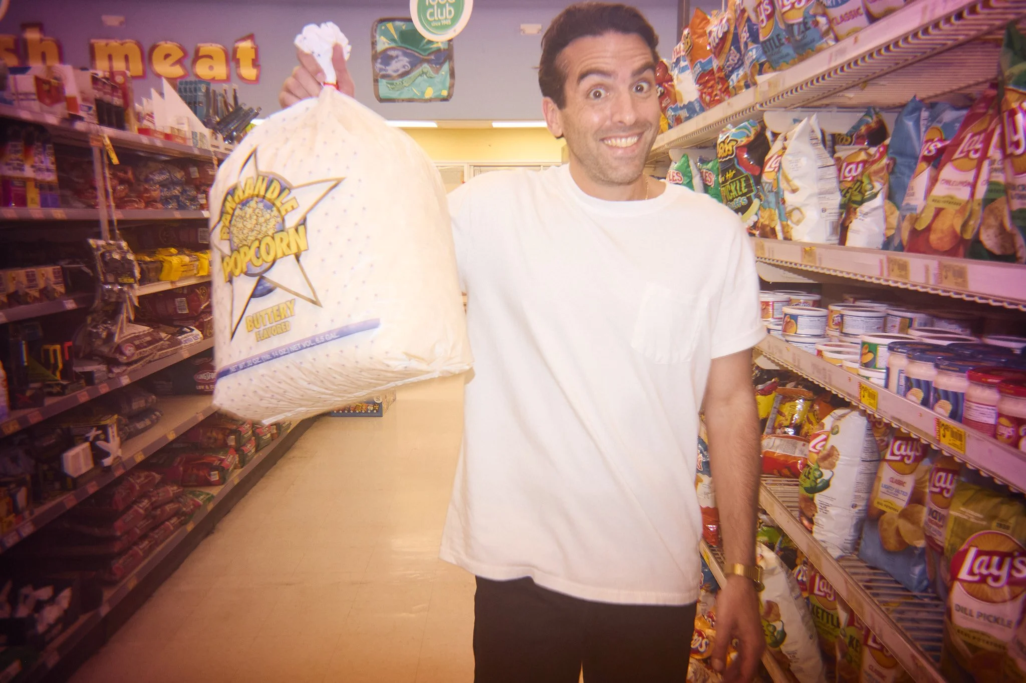 A happy man in a white T-shirt holding a large bag of popcorn in a grocery store aisle with snack shelves.