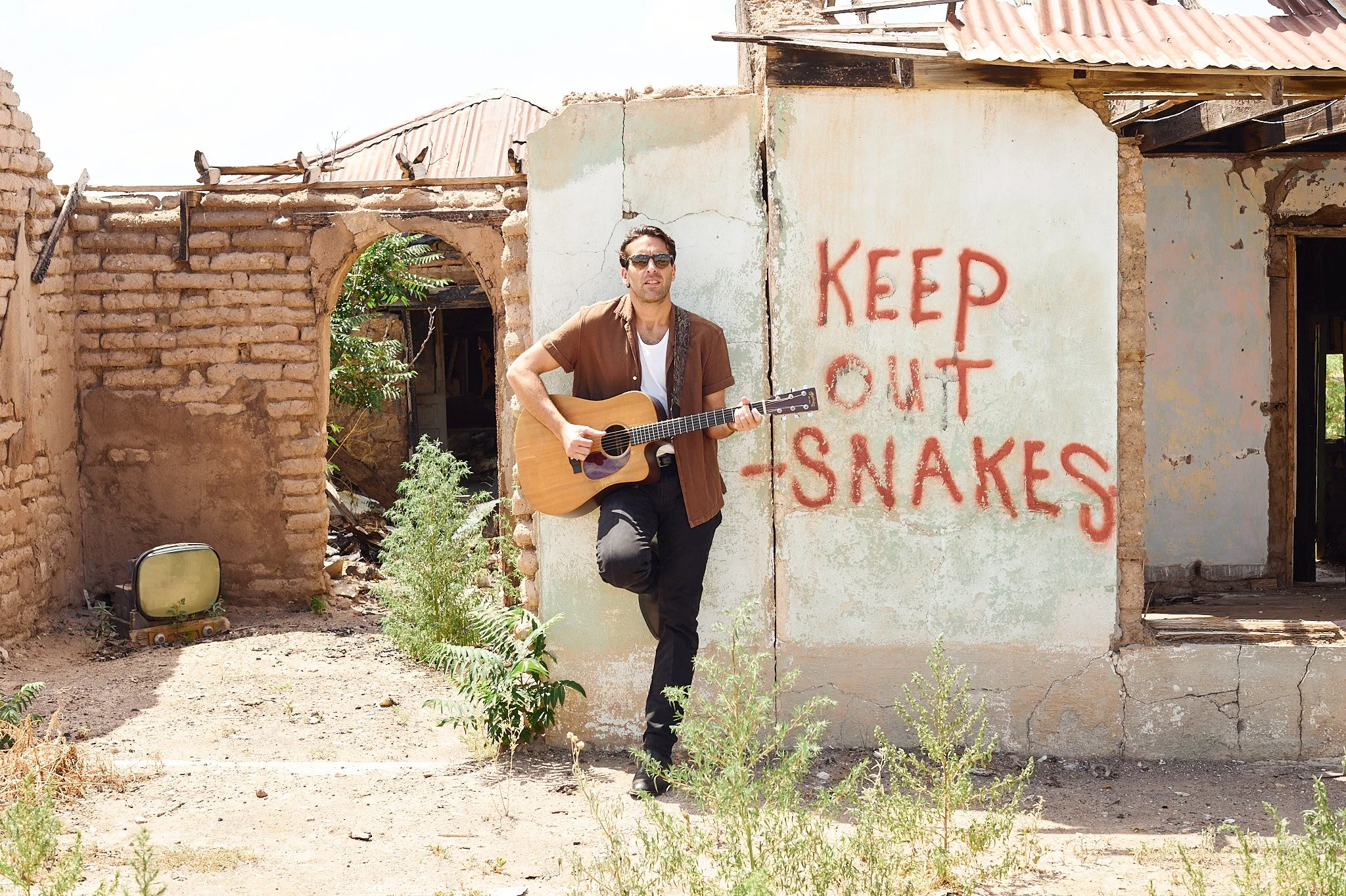 A man with sunglasses, wearing a brown shirt and black pants, standing with one foot raised against a weathered wall that has a red spray-painted sign saying "KEEP OUT SNAKES." The man is holding an acoustic guitar. The surroundings include an old, partially collapsed building with exposed bricks, wooden beams, and overgrown plants.