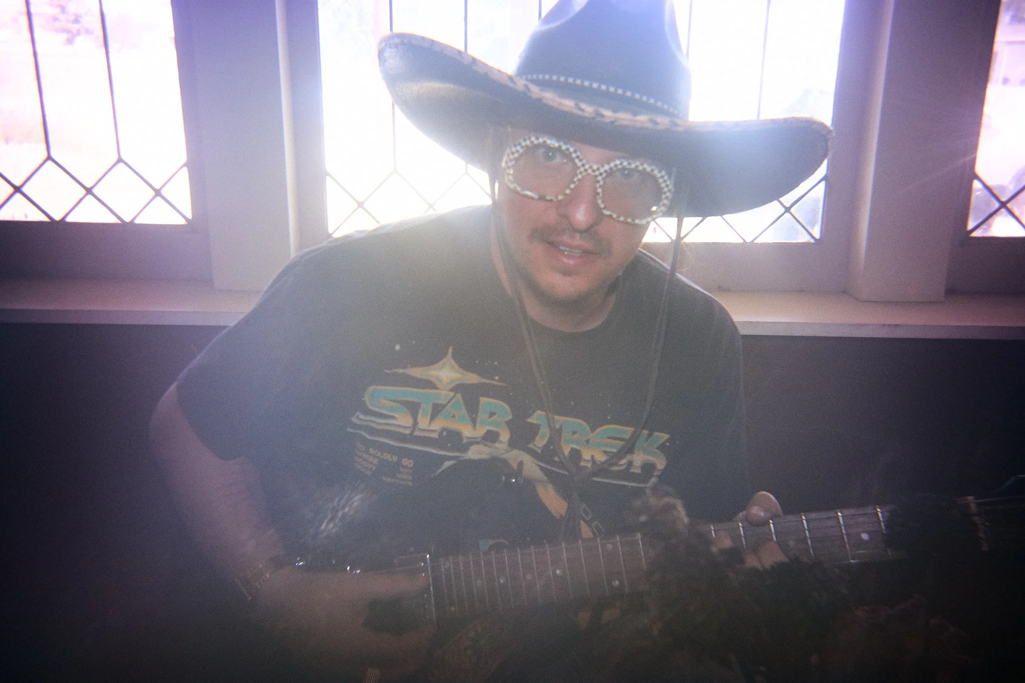 Person wearing a large cowboy hat and Star Wars T-shirt, sitting with a guitar, in front of a window with a grid pattern.