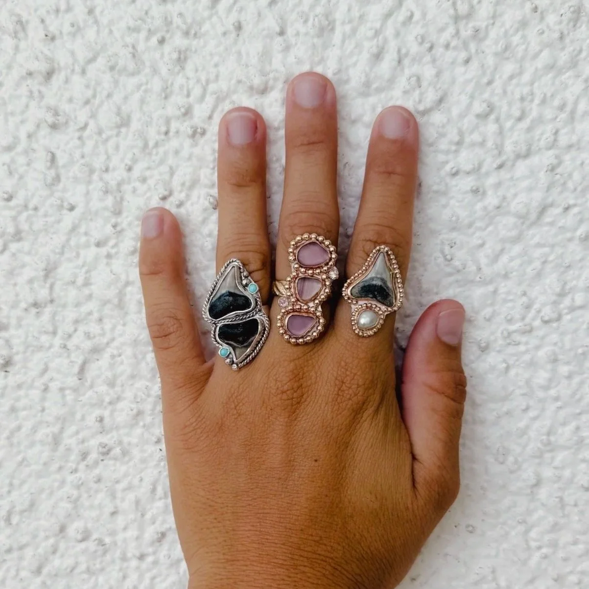 A hand against a white textured wall displaying three ornate rings with black, pink, and white stones.