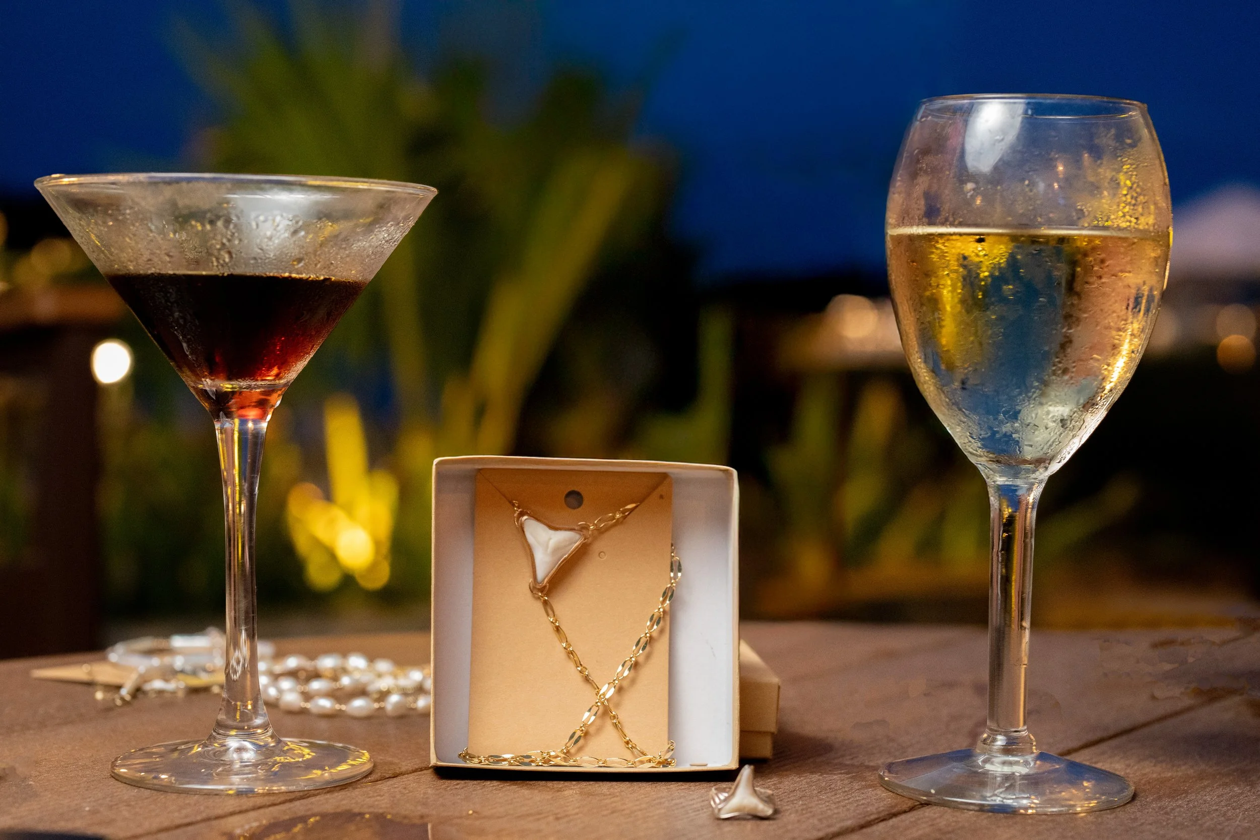 Two glasses of champagne, jewelry including a necklace and bracelet displayed on a small stand, with blurred background of plants and lights.