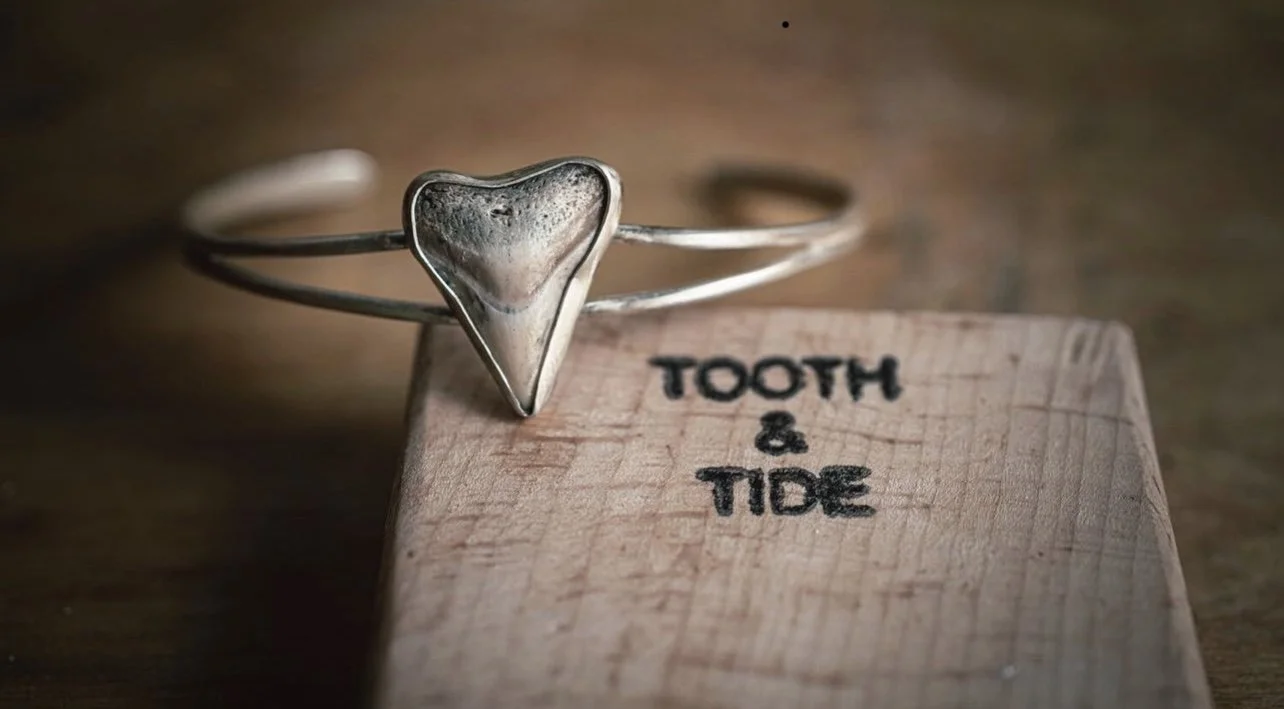 A silver bracelet with a heart-shaped design resting on a wooden surface next to a wooden block that reads 'TOOTH & TIDE'.