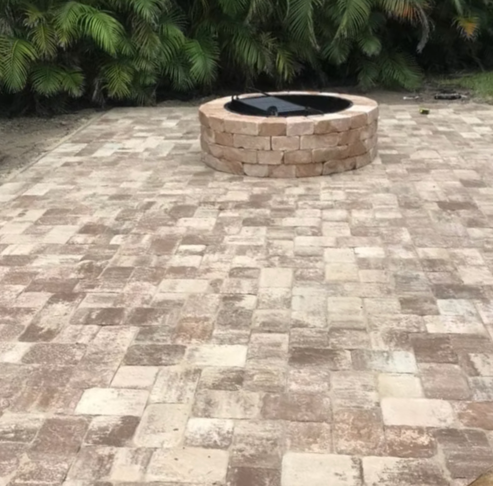 A brick fire pit with a black grill top on a paved patio with green shrubbery in the background.