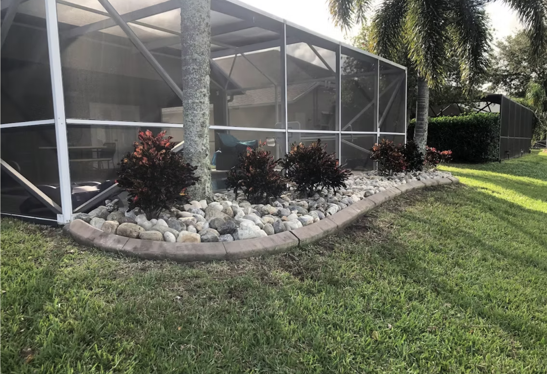 A backyard with a screened patio structure, surrounded by a landscaped garden bed with red-leafed plants, gray rocks, and a curved brick border, with green grass and tall trees in the background.