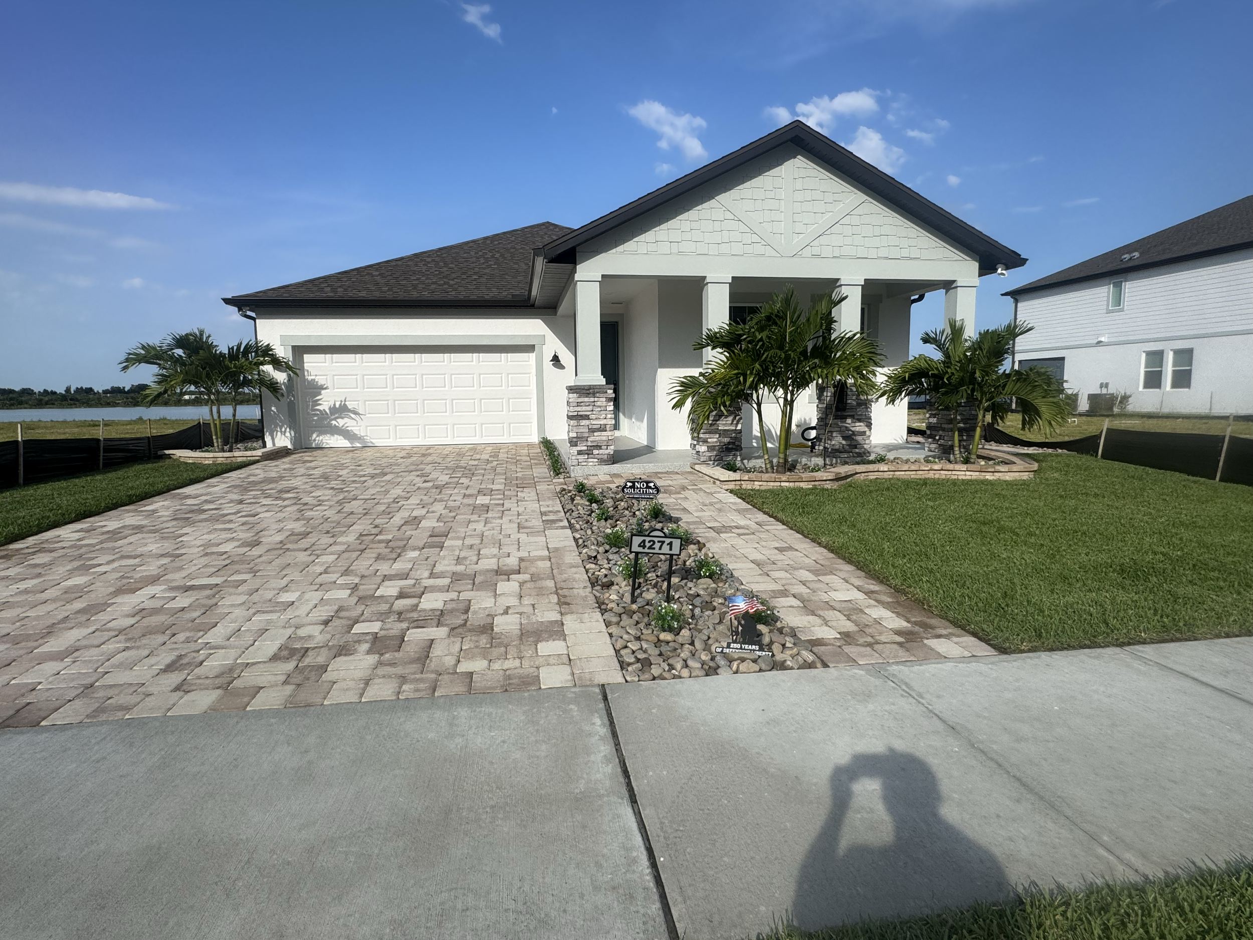 Front view of a modern white house with a grey shingled roof, driveway, and landscaped yard with palm trees. There are signs in the yard commemorating military service and a house number 4271.