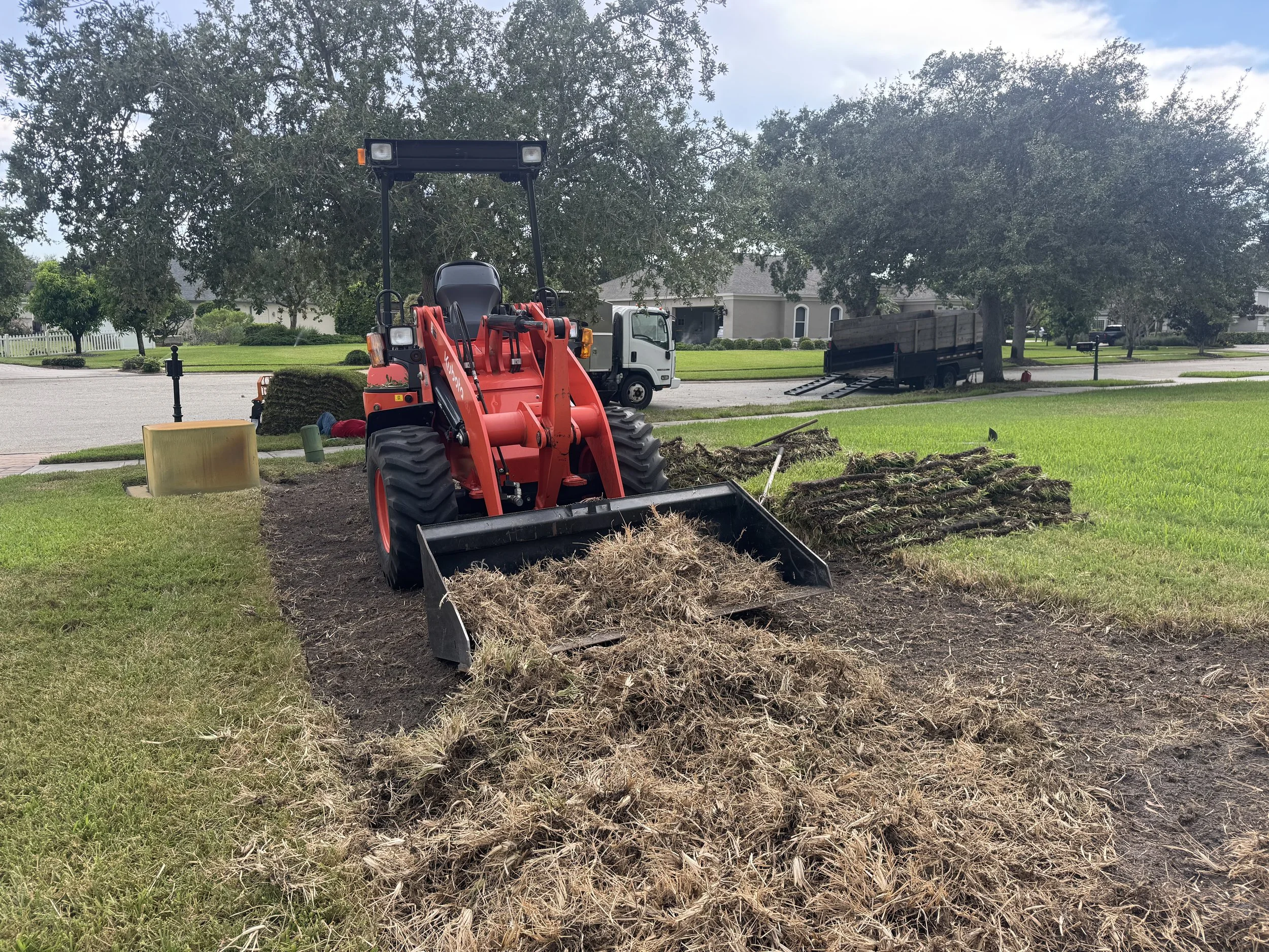 A front-end loader with red arms and black bucket scooping dirt in a residential yard, with a truck in the background, large trees, houses, and a green lawn.