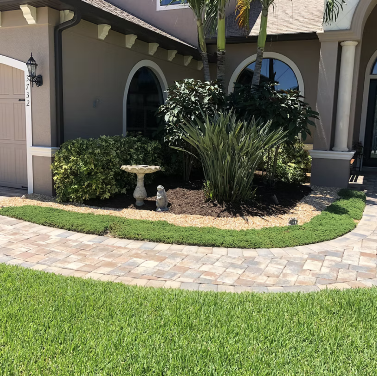 Front yard landscaping with green grass, a brick pathway, a landscaped flower bed with bushes and tall plants, and decorative garden statues in front of a house with arched windows and columns.