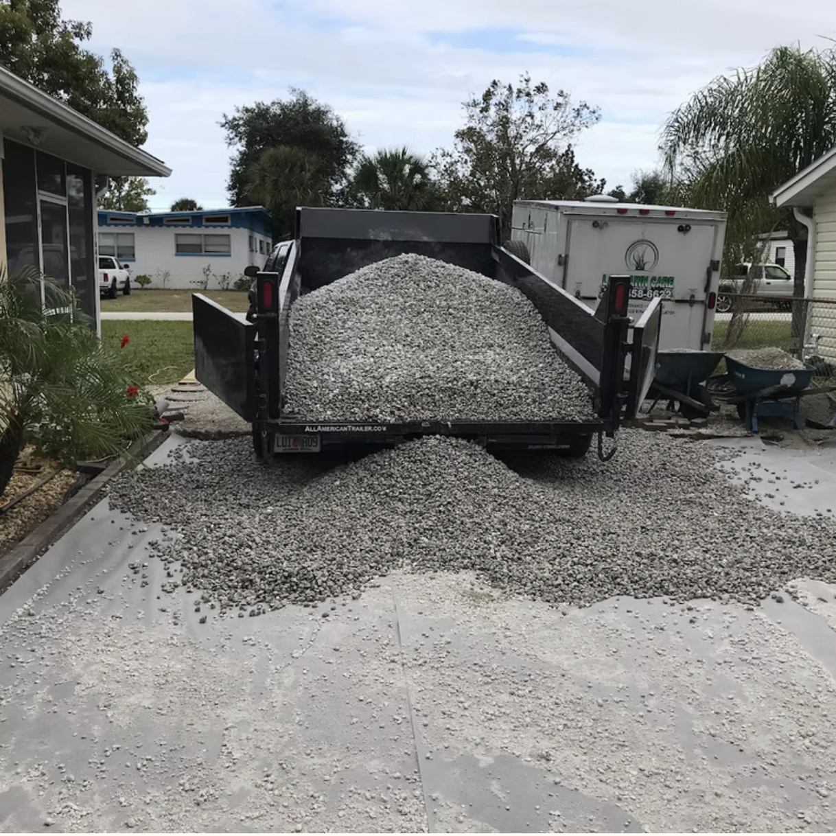 A black utility trailer filled with gray gravel, with some spilling onto a concrete driveway.