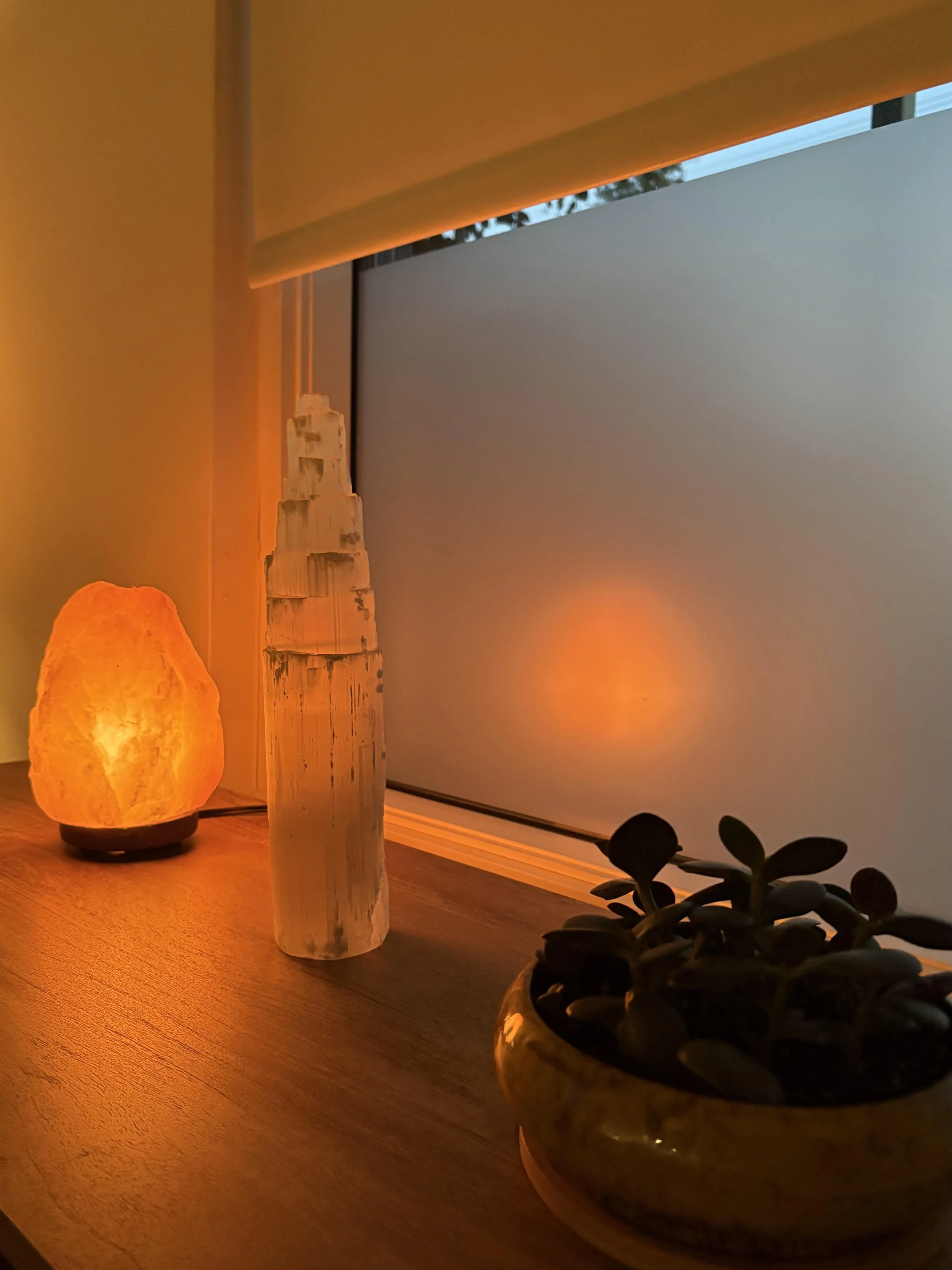 A warm-toned salt lamp and a small potted plant on a wooden surface, with a white candle holder and a white brick structure in the background, and a window with a view of trees outside.
