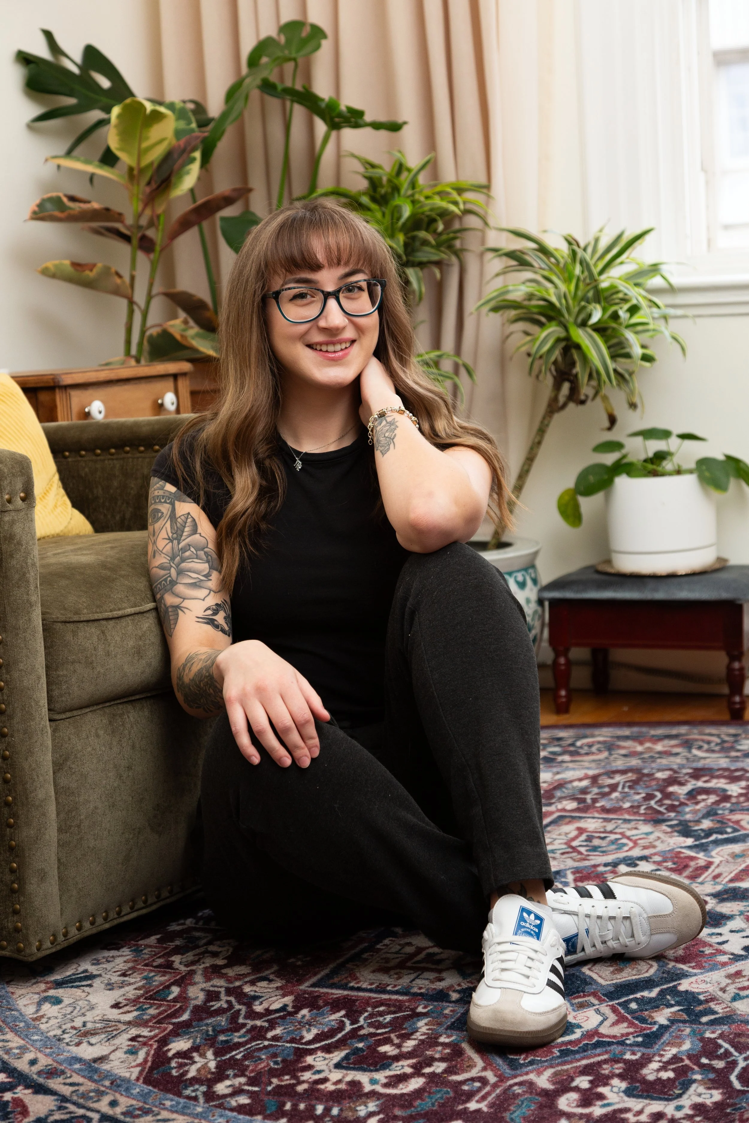 A young woman with long brown hair, glasses, and tattoos wearing a black t-shirt and black pants, sitting on the floor in a living room surrounded by houseplants, with a wooden couch and an oriental rug in the background.