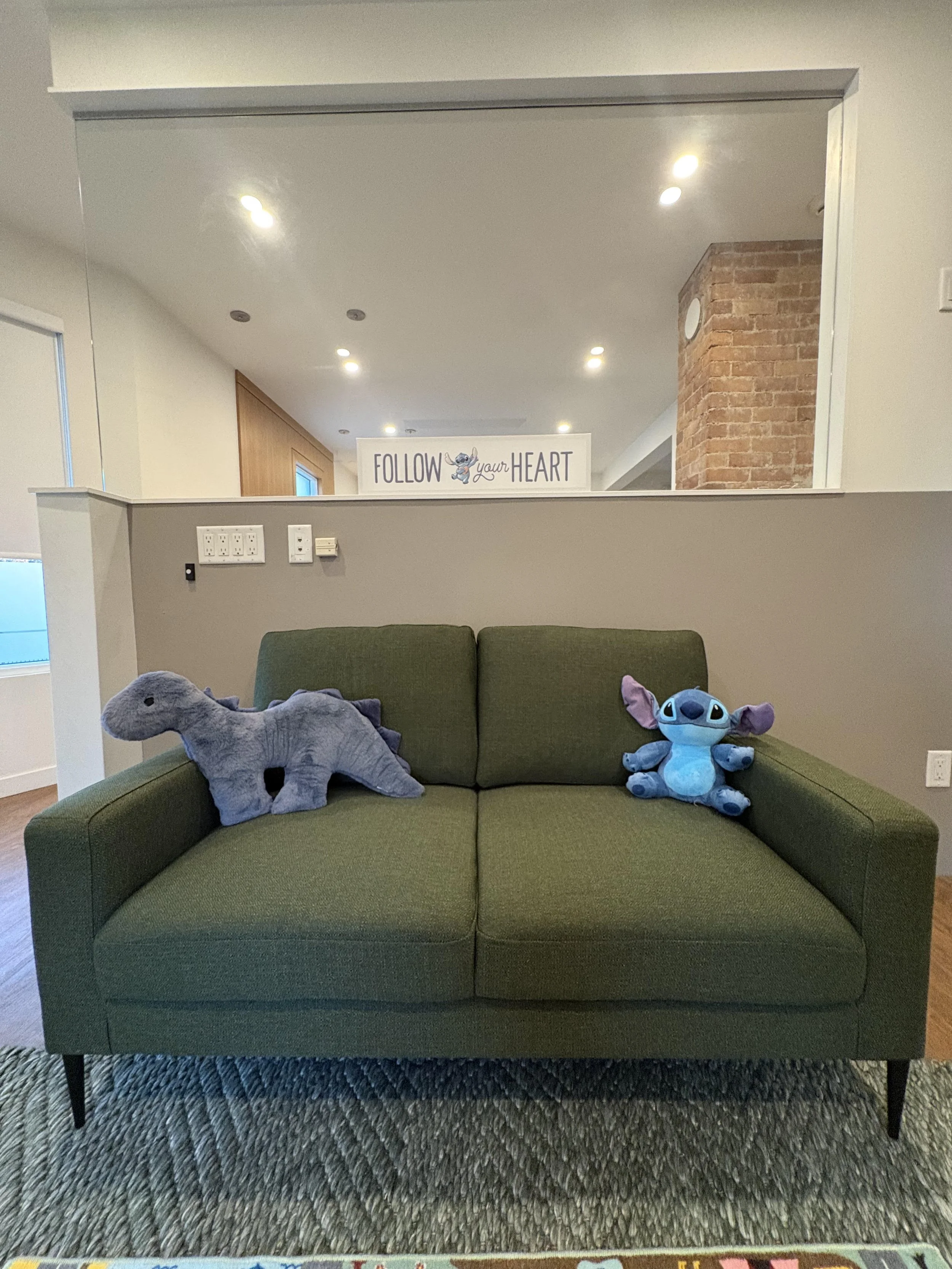 A green sofa with two plush toys, a gray dinosaur and a blue creature, sitting on it in a living room with a sign that reads 'Follow your Heart' in the background.